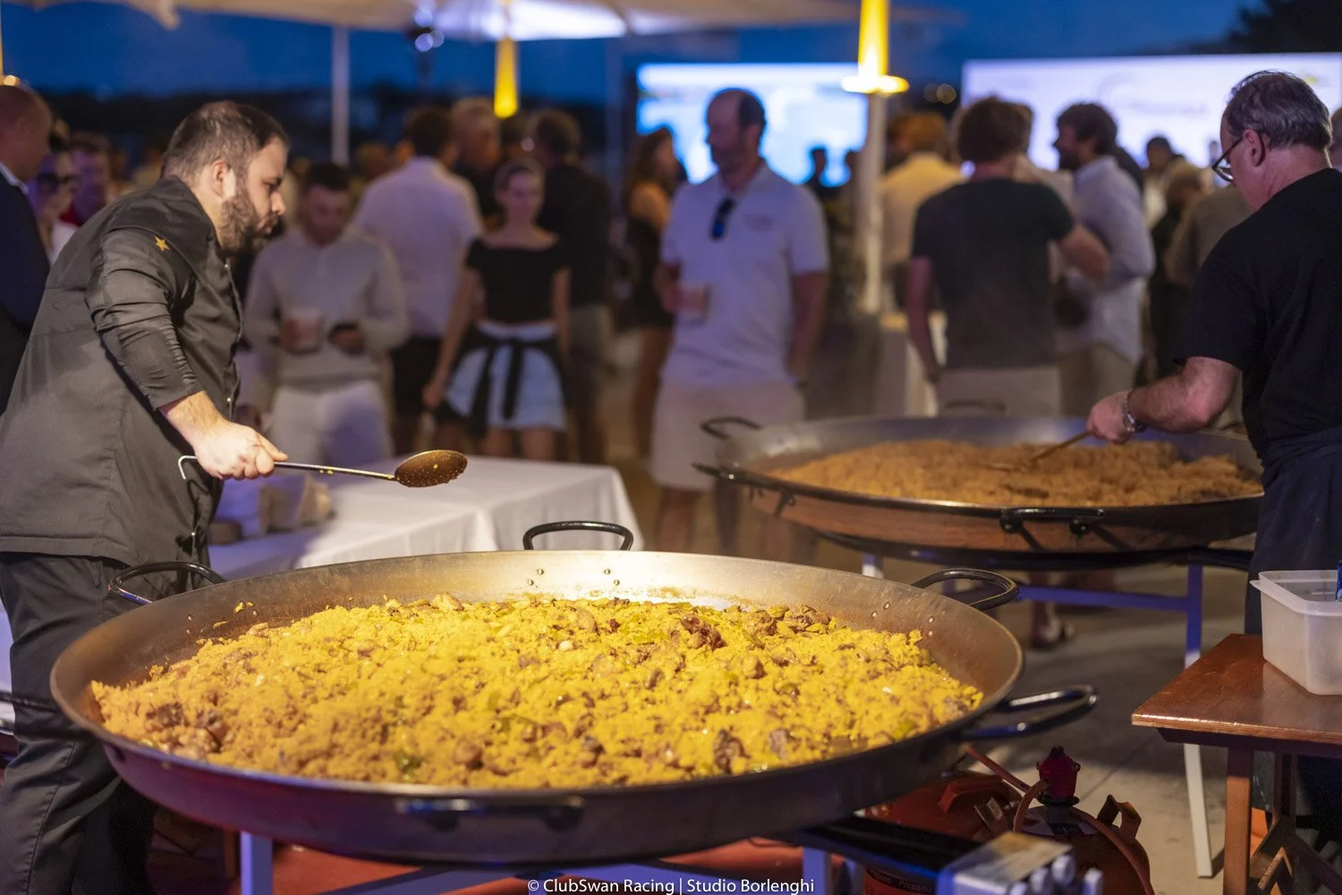 Dos cocineros preparando paella en un evento con muchas personas en un ambiente social y de comida.