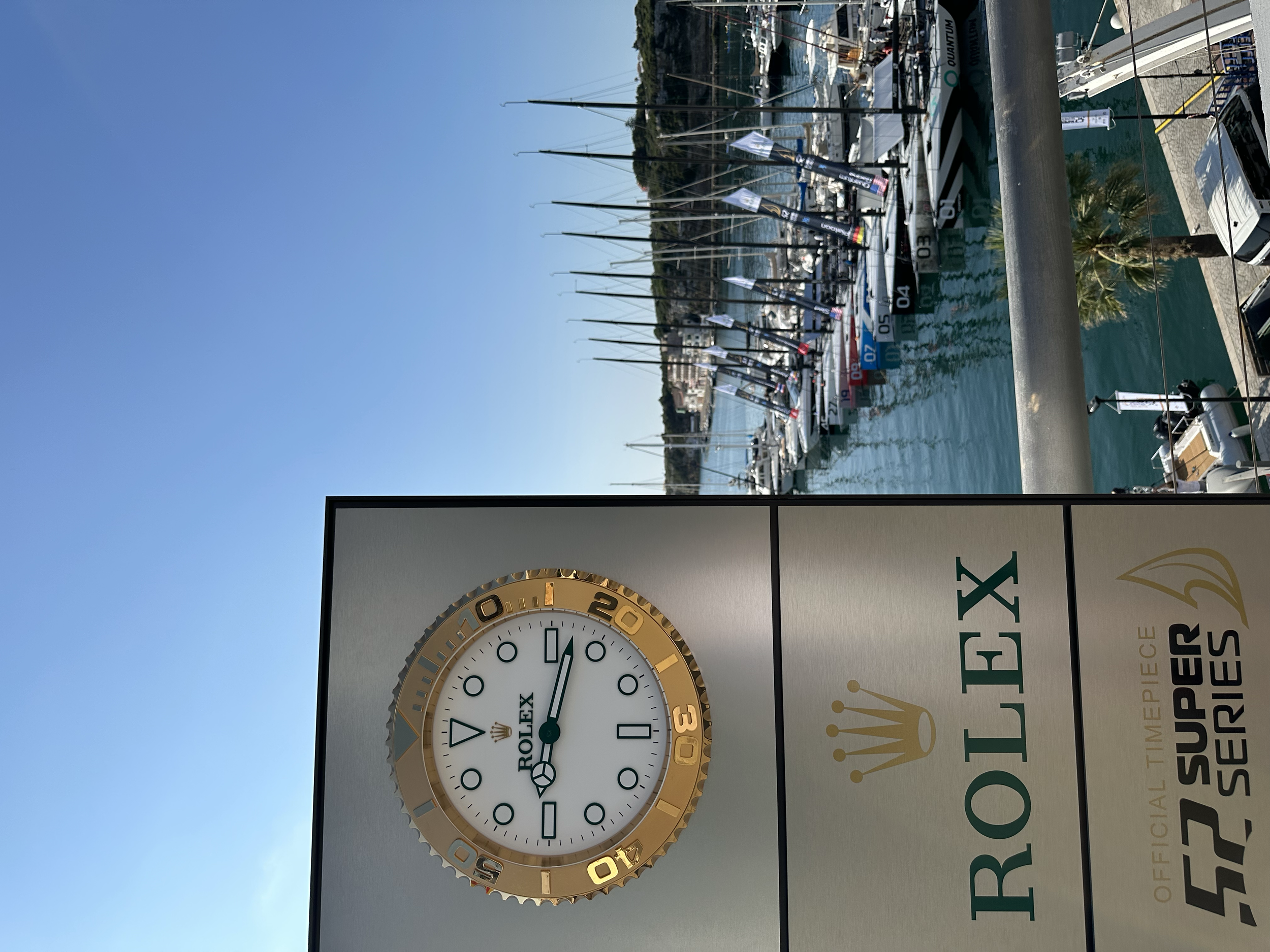 Cartel de Rolex con reloj de cuerda automática, lado izquierdo vista del puerto con yates y barcos en el agua, cielo despejado.