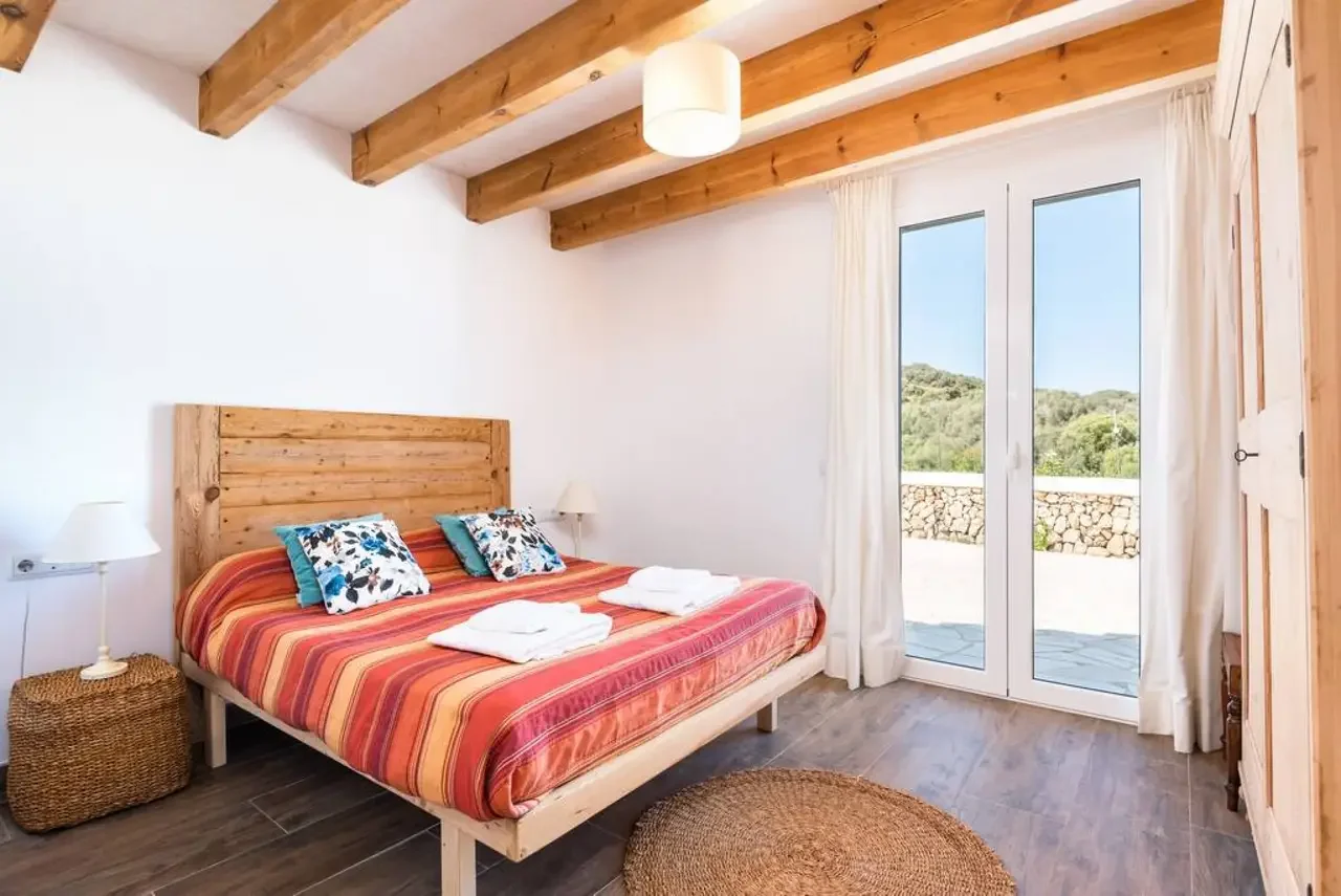 Habitación con cama, cabecera de madera, ventanas de vidrio con cortinas blancas, vista a naturaleza, suelo de madera, lámparas de noche, alfombra de fibra, techo con vigas de madera.