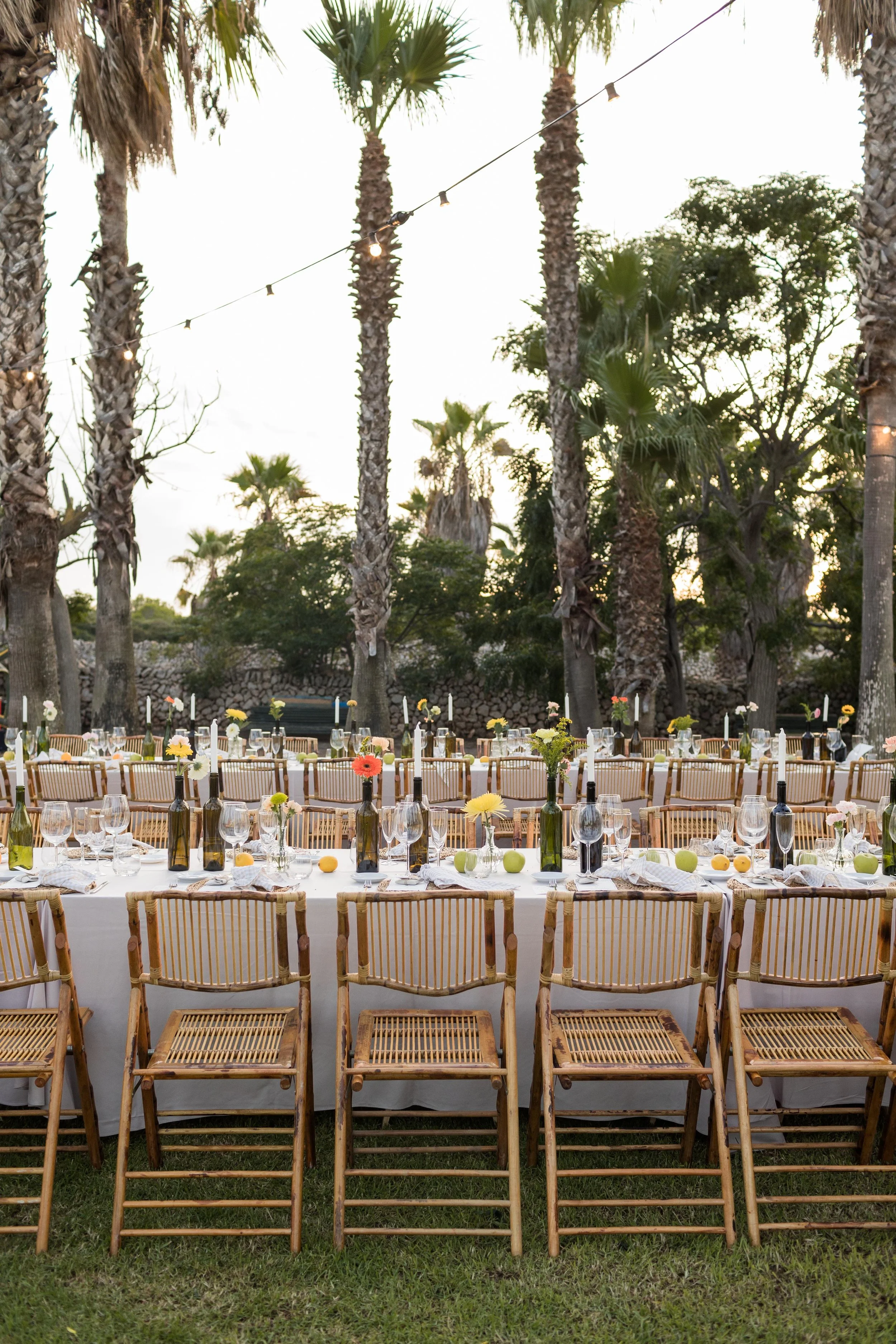Mesas decoradas al aire libre con sillas de madera, botellas con flores, copas y platillos, rodeadas de palmeras y árboles, con luces colgantes, en un ambiente de jardín al atardecer.