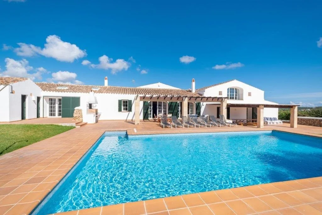 Casa blanca con piscina y tumbonas bajo un cielo azul con algunas nubes.
