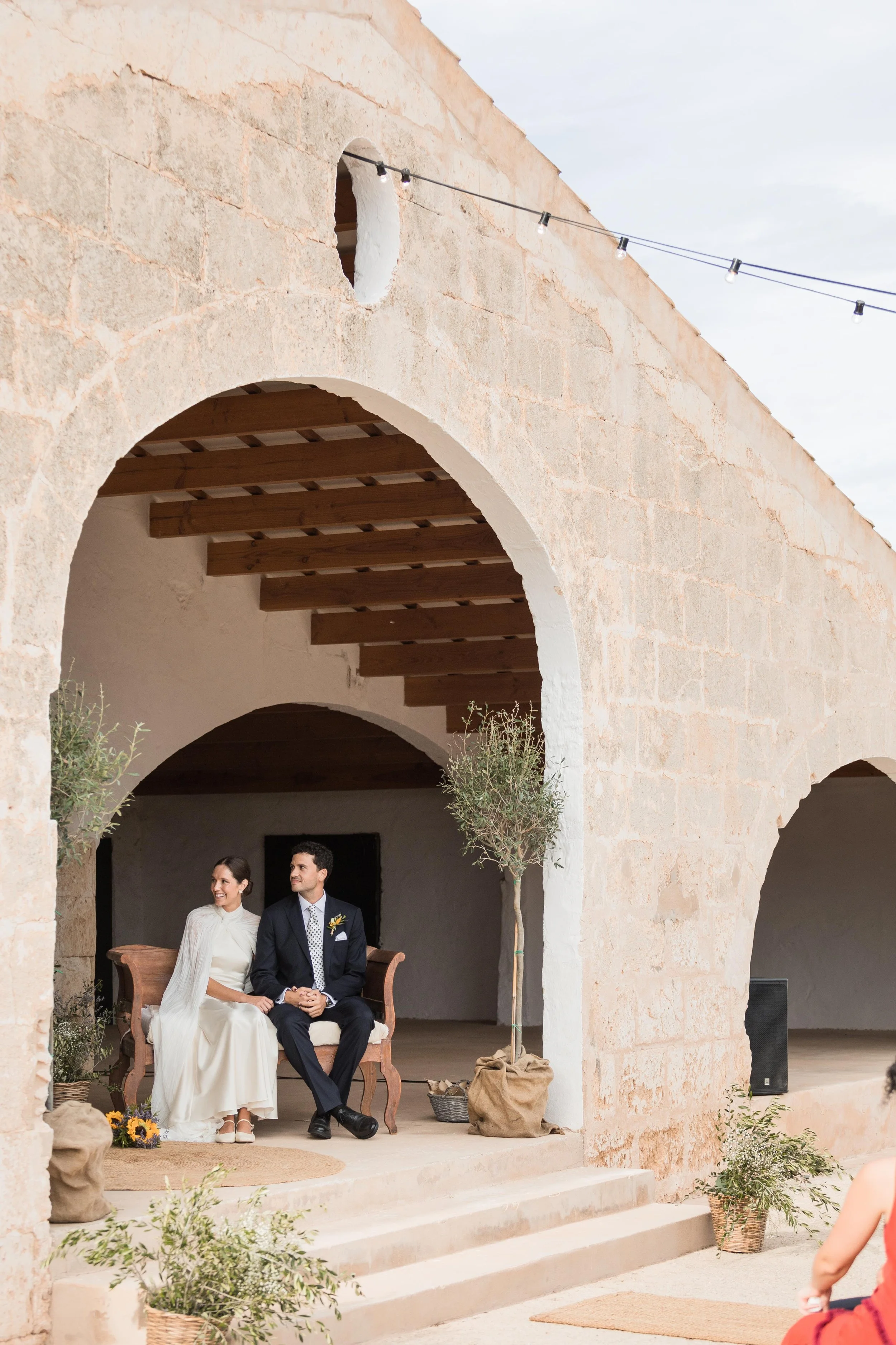 Pareja de novios en boda sentados en un banco en un espacio al aire libre con arcos de piedra y decoraciones de plantas y arena.
