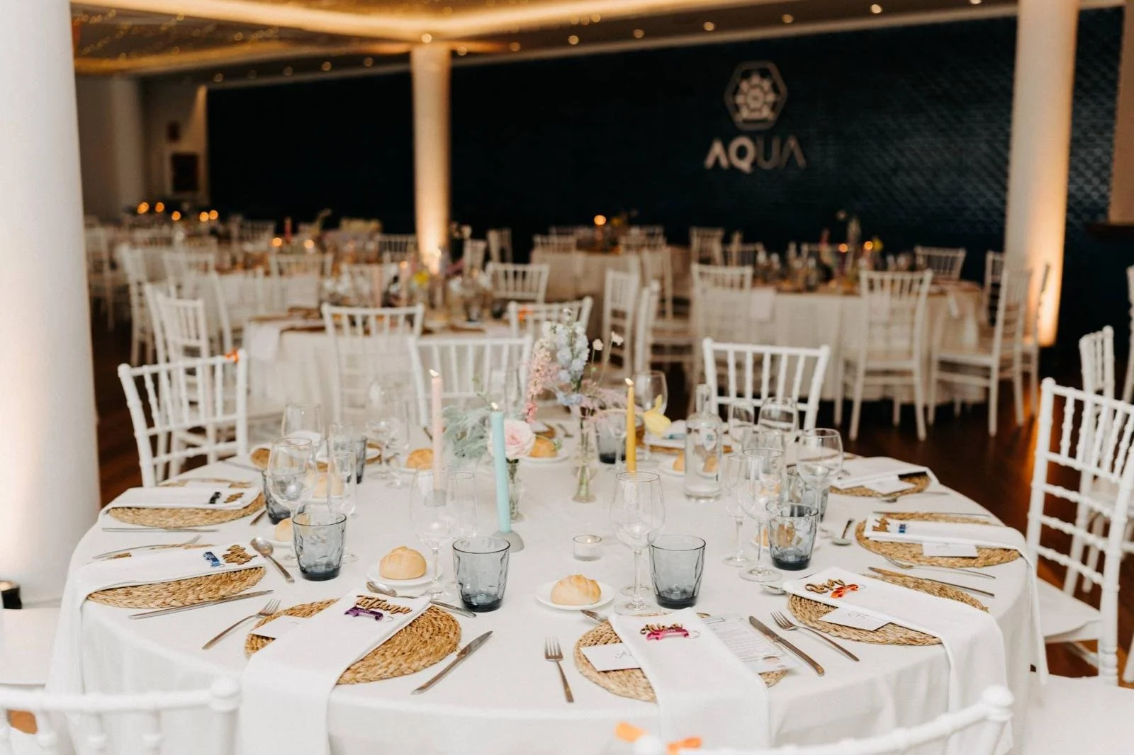Mesa decorada para evento con platos, cubiertos, copas, jarras de agua, velas y centros de mesa con flores en un salón con paredes negras y decoraciones elegantes.