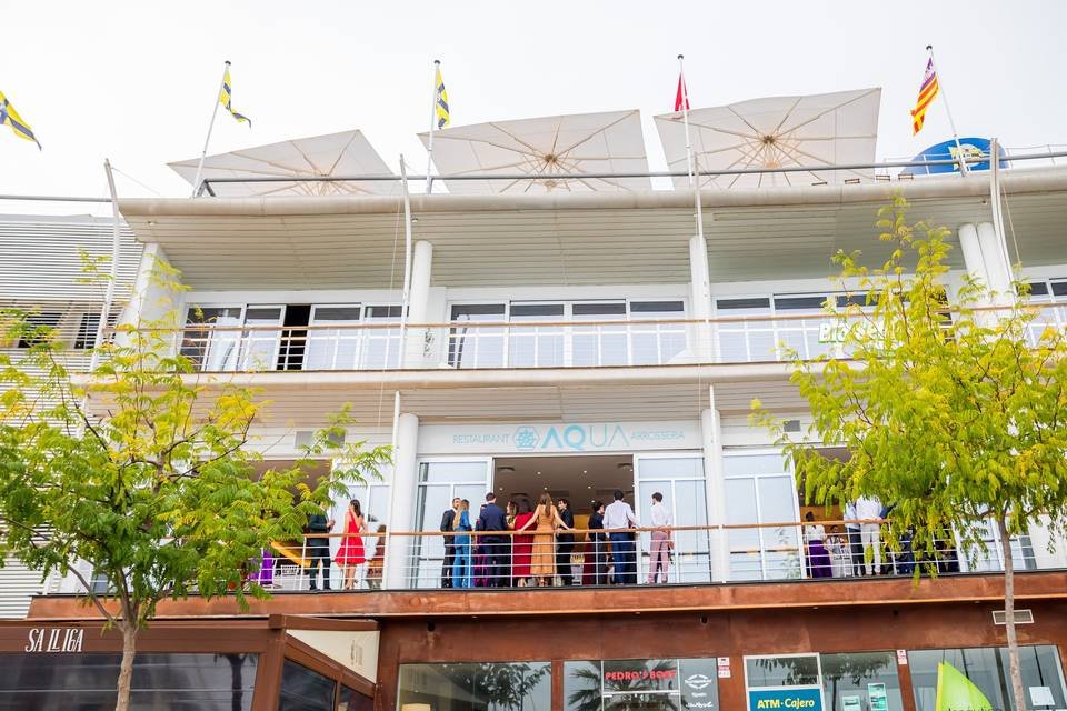 Edificio de varios pisos con una colección de personas en la entrada principal, con cartel que dice 'Restaurante Aqua' y varios paraguas en la terraza, rodeado de árboles verdes.