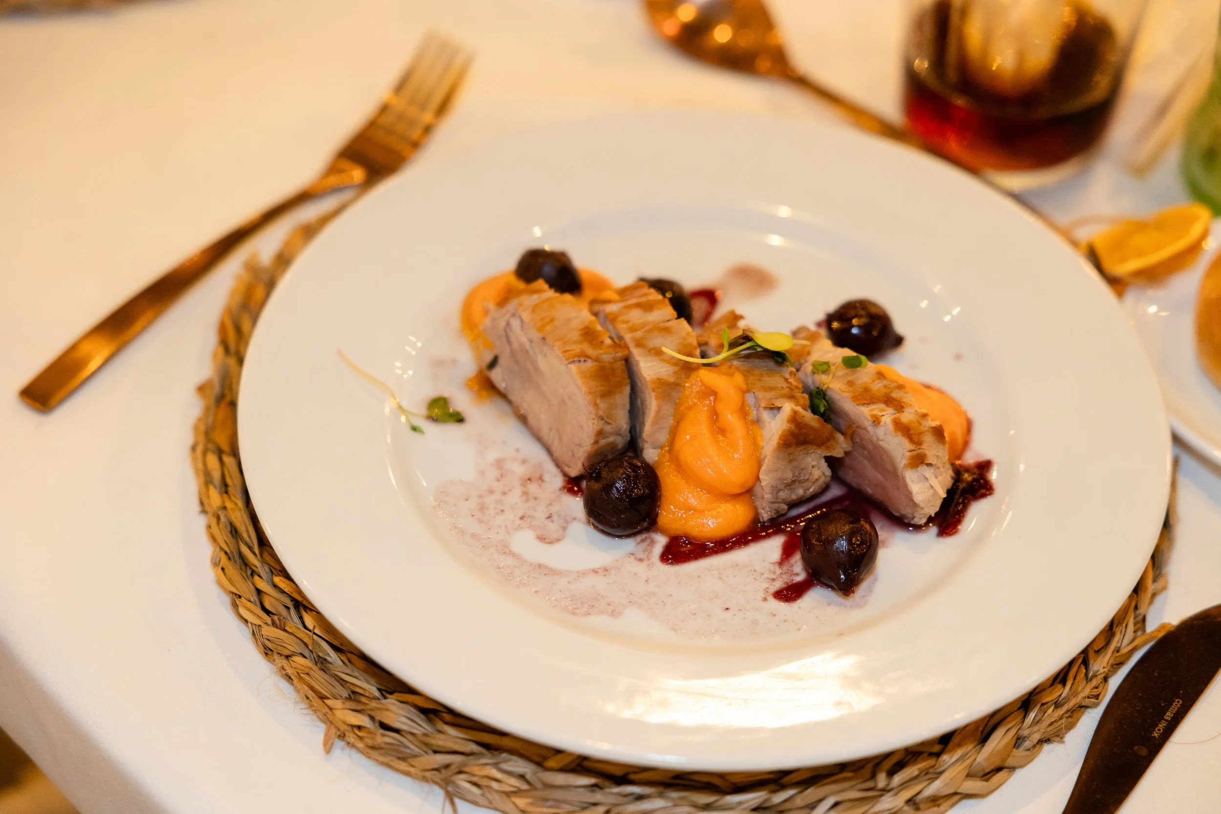Plato de carne con puré de papas, aceitunas negras y salsa en un plato blanco sobre un tapete de mimbre en una mesa con cubiertos de oro