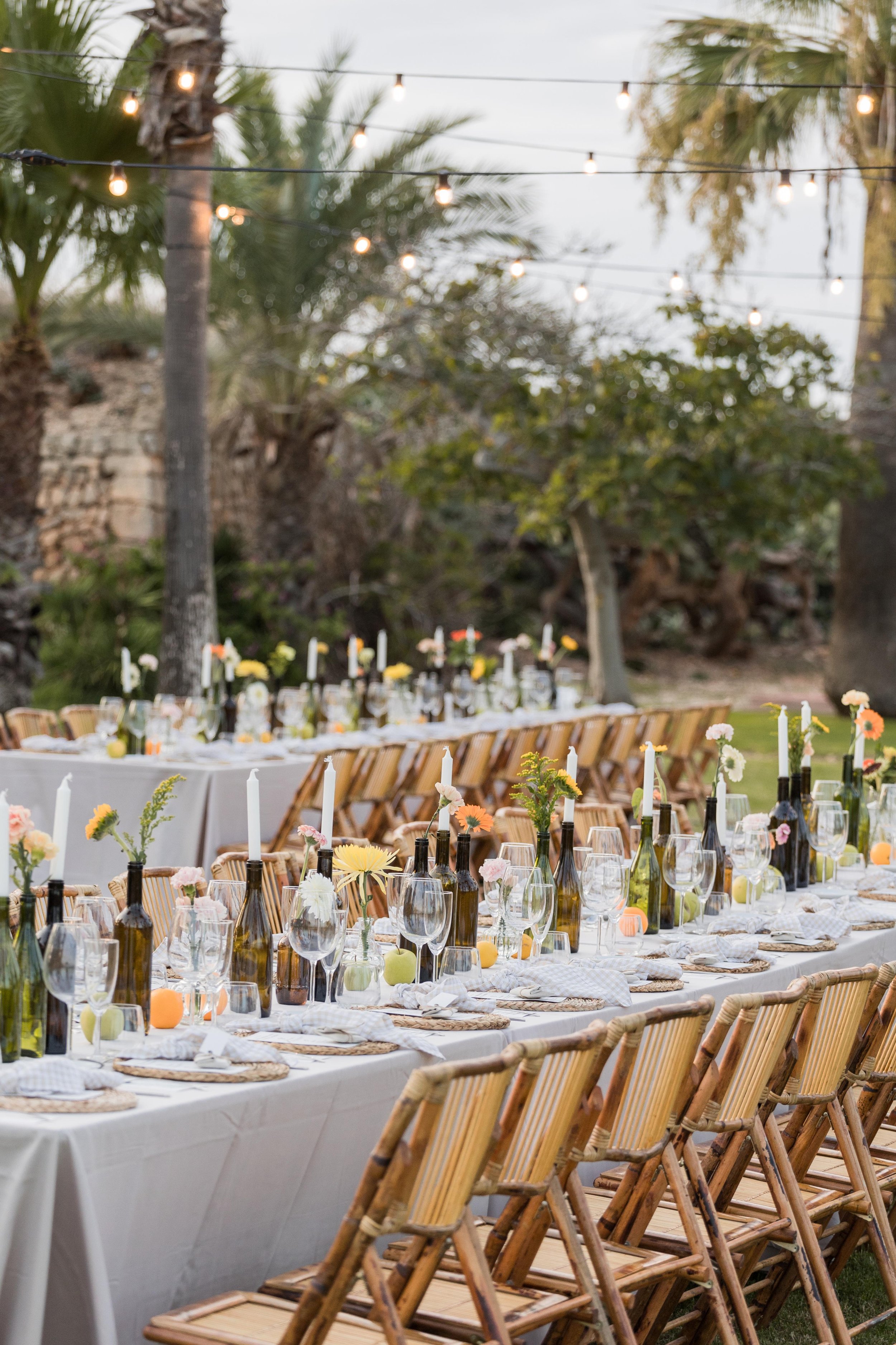 Decoración de una cena al aire libre con mesas largas, sillas de madera, velas en botellas, flores y luces colgantes, en un ambiente natural con árboles y plantas.