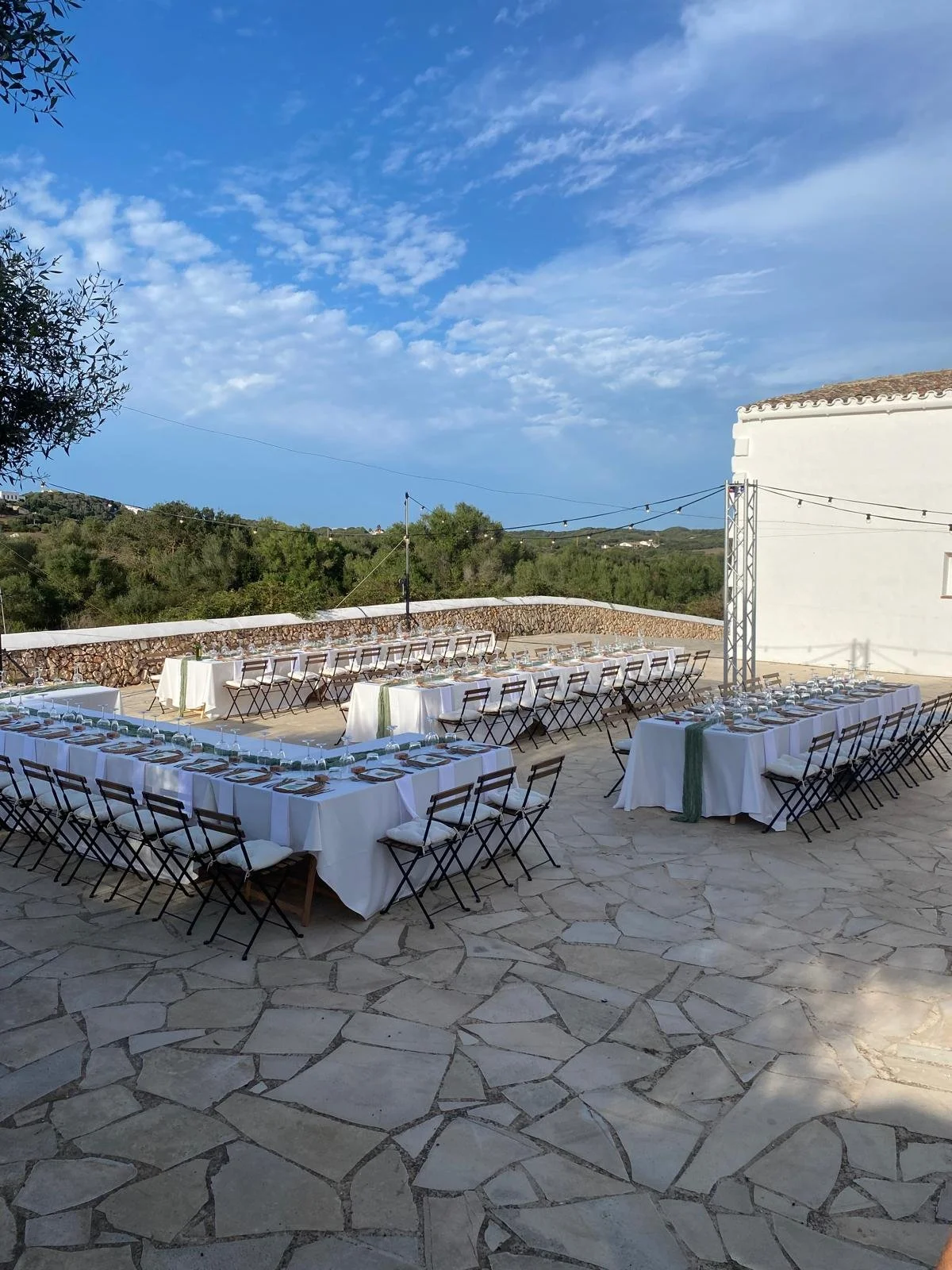 Mesa larga decorada con manteles blancos y vajilla, dispuesta para una celebración al aire libre en un patio con vista a montañas y cielo despejado.