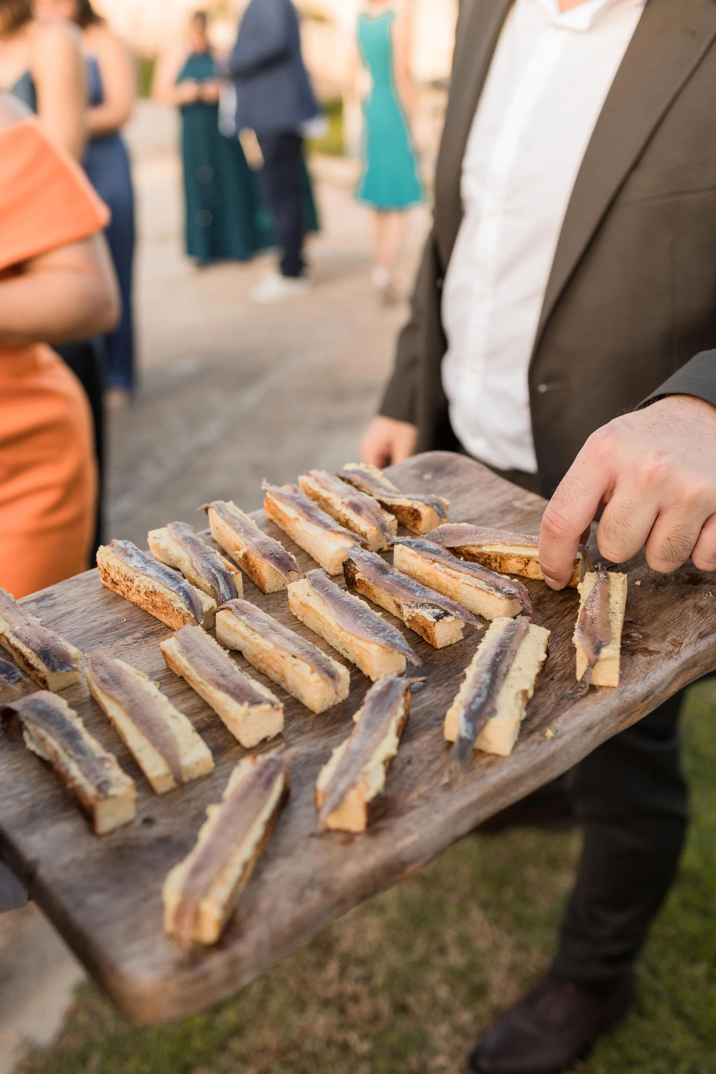 Persona sirviendo bocados de anchoa sobre una tabla de madera en una reunión al aire libre.