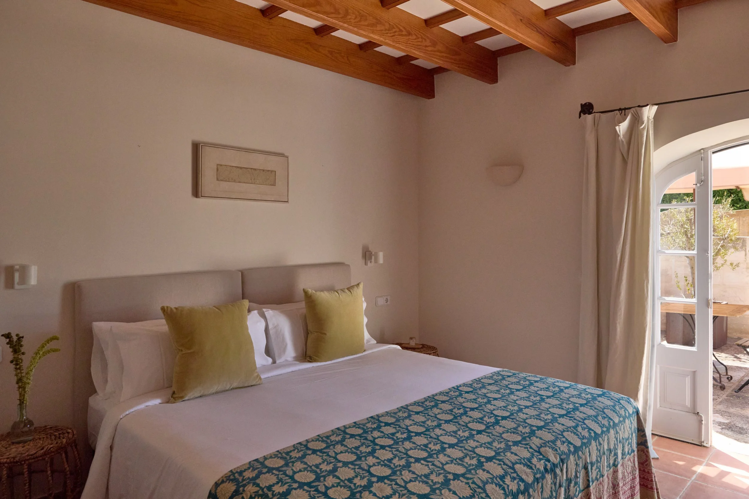 Habitación con cama doble, cojines amarillos, cortinas blancas y puerta de cristal que da a una terraza exterior.