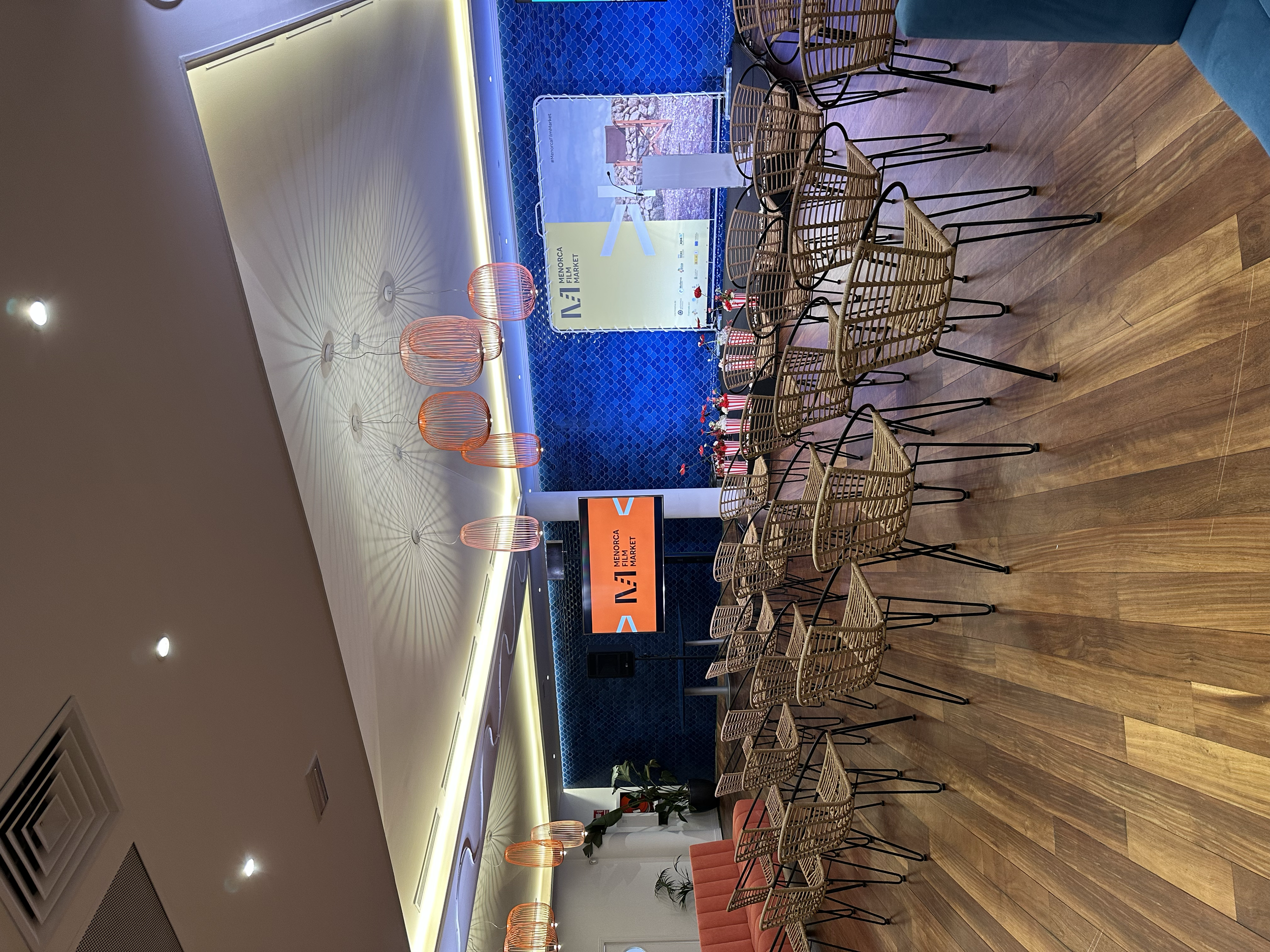 Sala de eventos con mesas y sillas de mimbre, decoraciones modernas y una pantalla digital con logotipo, iluminación de techo y paredes azules.