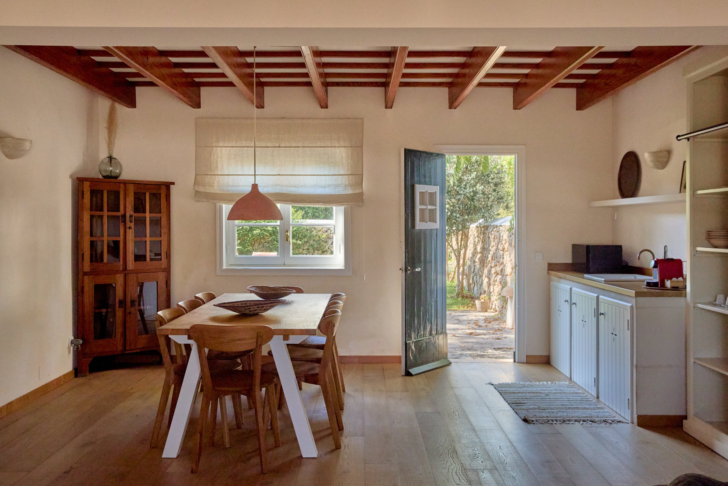 Interior de una casa con mesa de comedor de madera, armario de madera, puerta abierta hacia un jardín exterior, y ventanas con cortinas claras.