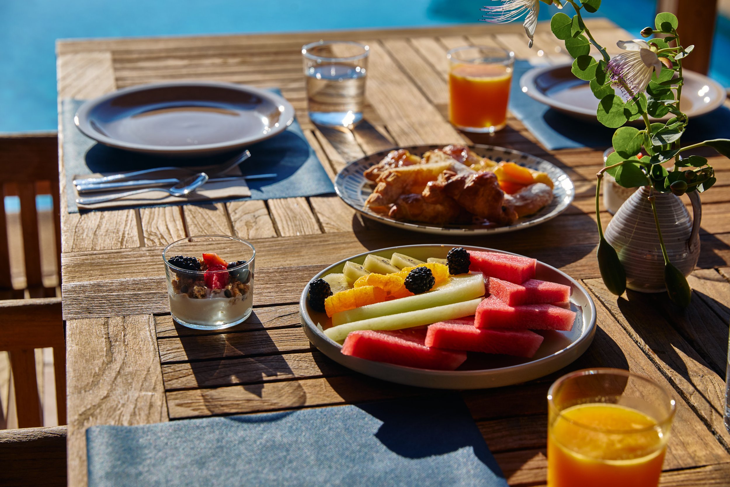 Mesa de desayuno con frutas, jugo de naranja, yogur con berries, pollo frito, y otros alimentos, en un entorno con luz natural y flores decorativas.