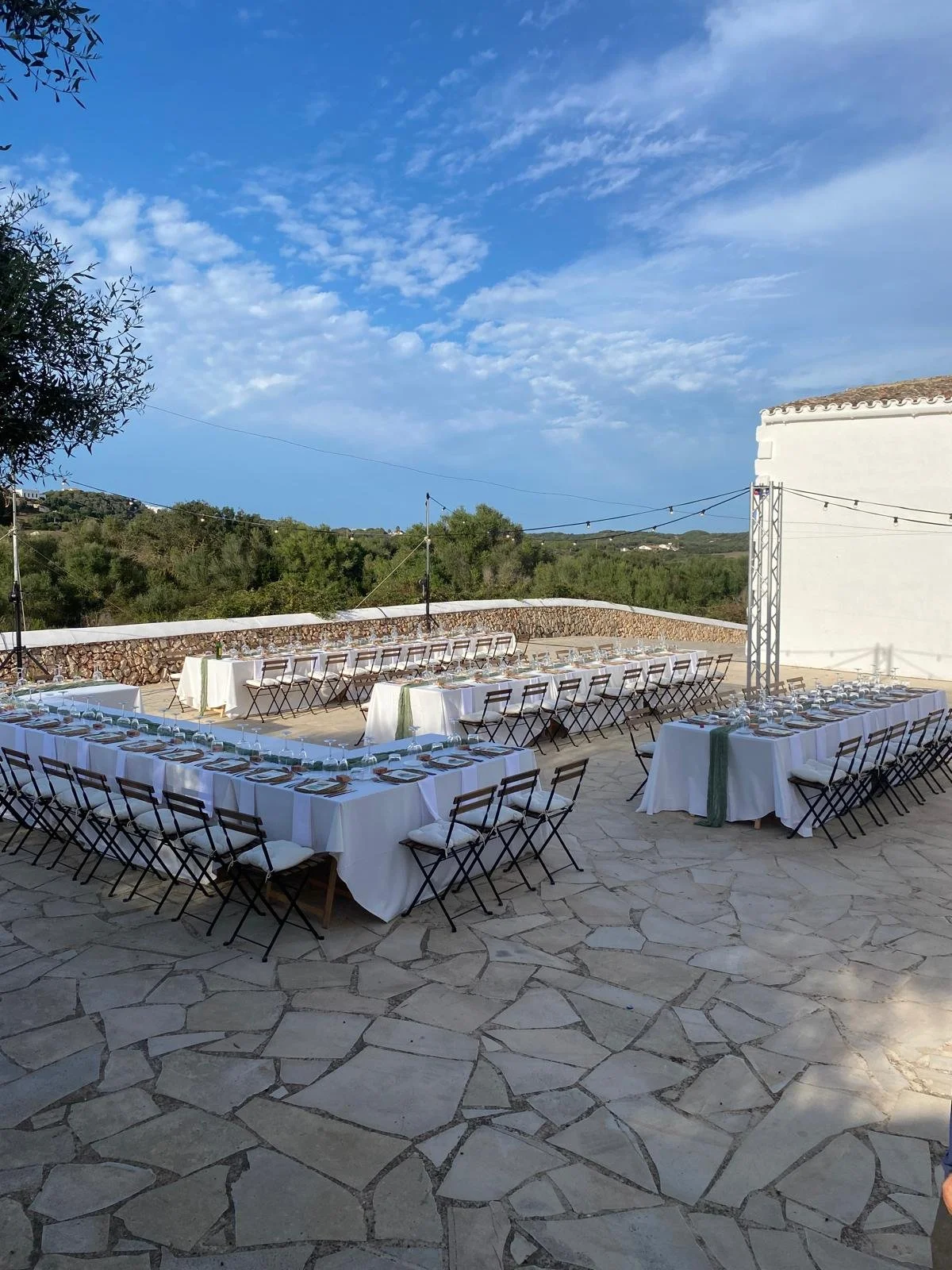 Una mesa larga decorada para una ceremonia o evento al aire libre, con manteles blancos, vajilla y cubiertos, en un espacio exterior con suelo de piedra y rodeado de vegetación, bajo un cielo azul con algunas nubes.