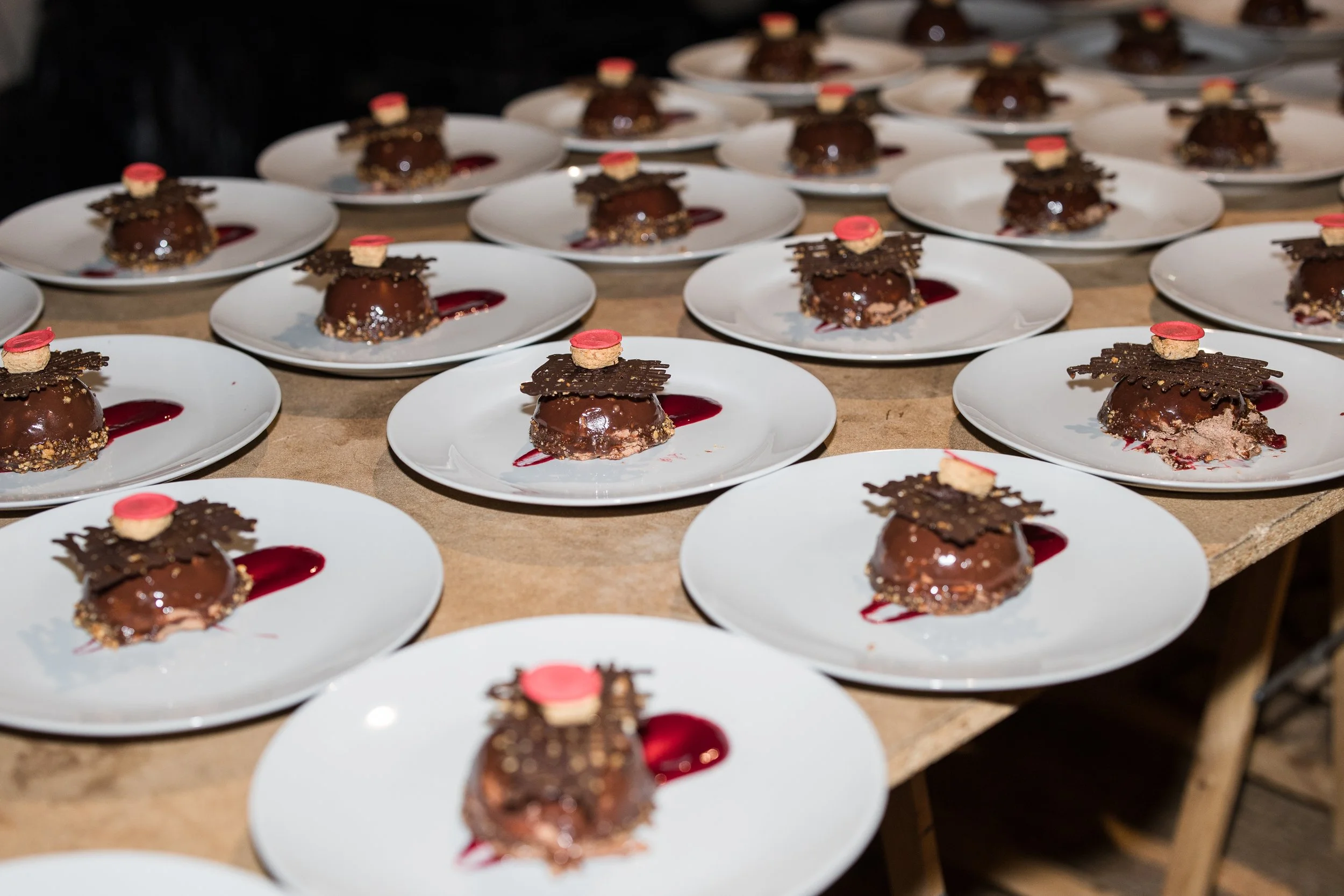 Varios postres de chocolate decorados con pequeños pastelitos rosados y con una salsa de fruta roja en platos blancos sobre una mesa de madera.
