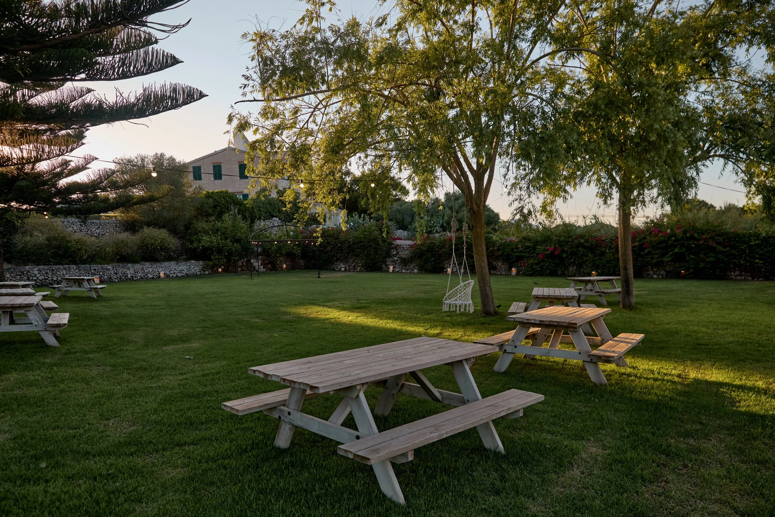 Patio con mesas de picnic de madera, columpio de cuerda colgado en un árbol, césped verde, árboles y flores en un fondo, luces colgantes, en un ambiente al atardecer en un jardín o parque.