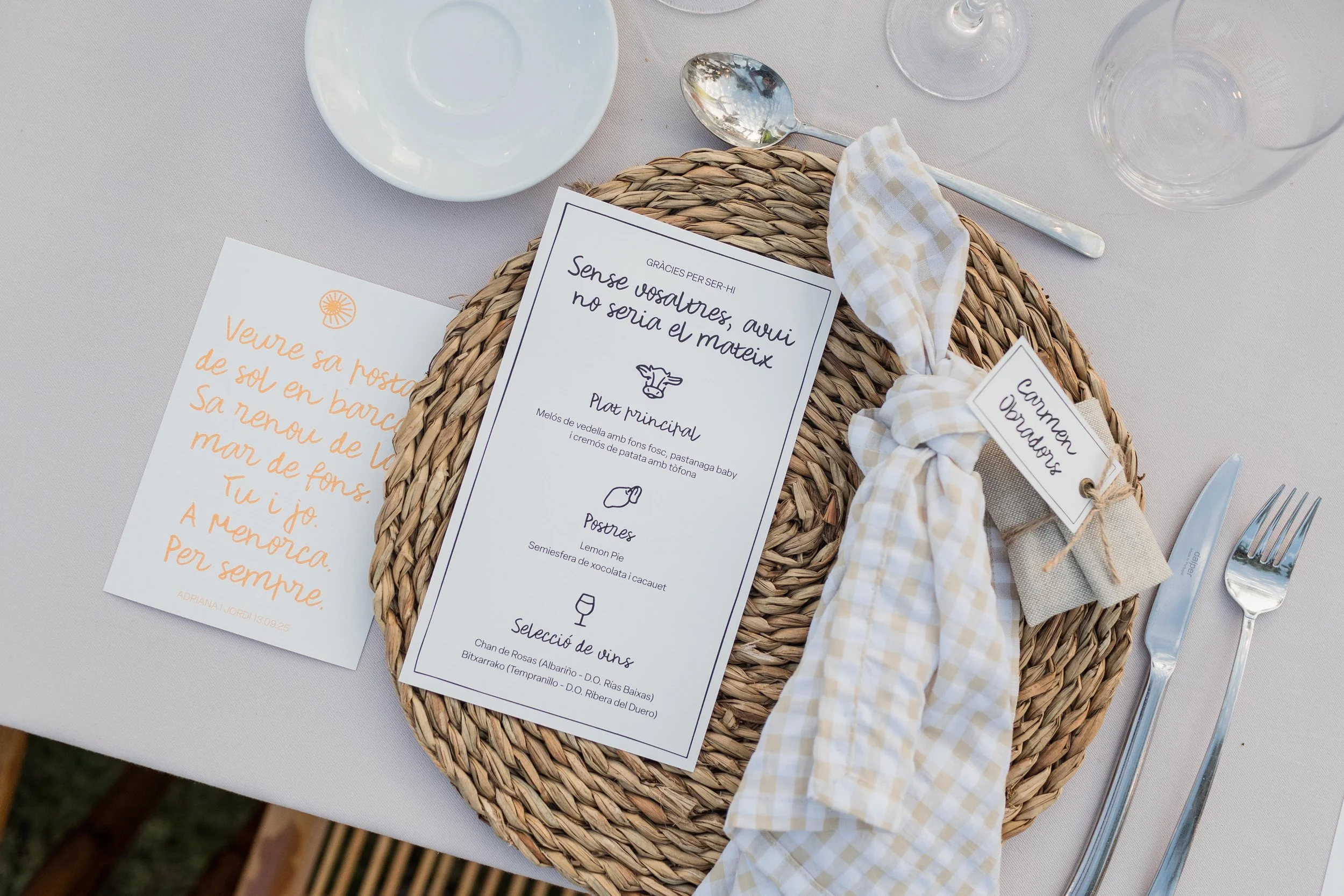 Mesa de boda con plato de mimbre, servilleta atada con cuerda, tarjeta con menú, cubiertos, vaso y copa, y nota escrita en papel color crema con letras naranja.