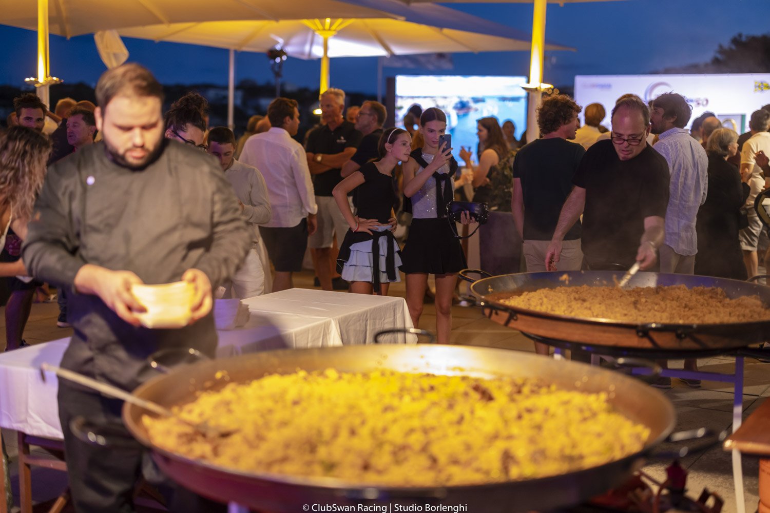 Persona sirviendo comida en un evento nocturno con muchas personas y puestos de comida.