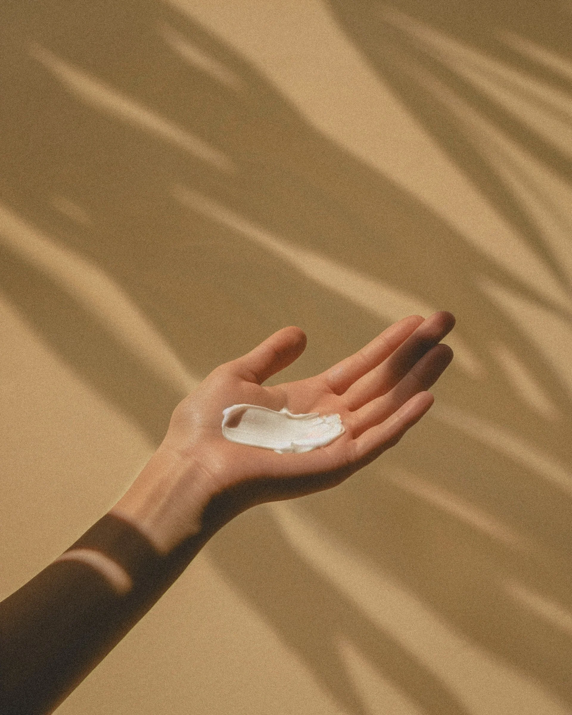 Eine Hand mit weißer Creme auf der Handfläche, Schatten eines Zimmerpflanzenblattes auf der Wand im Hintergrund.
