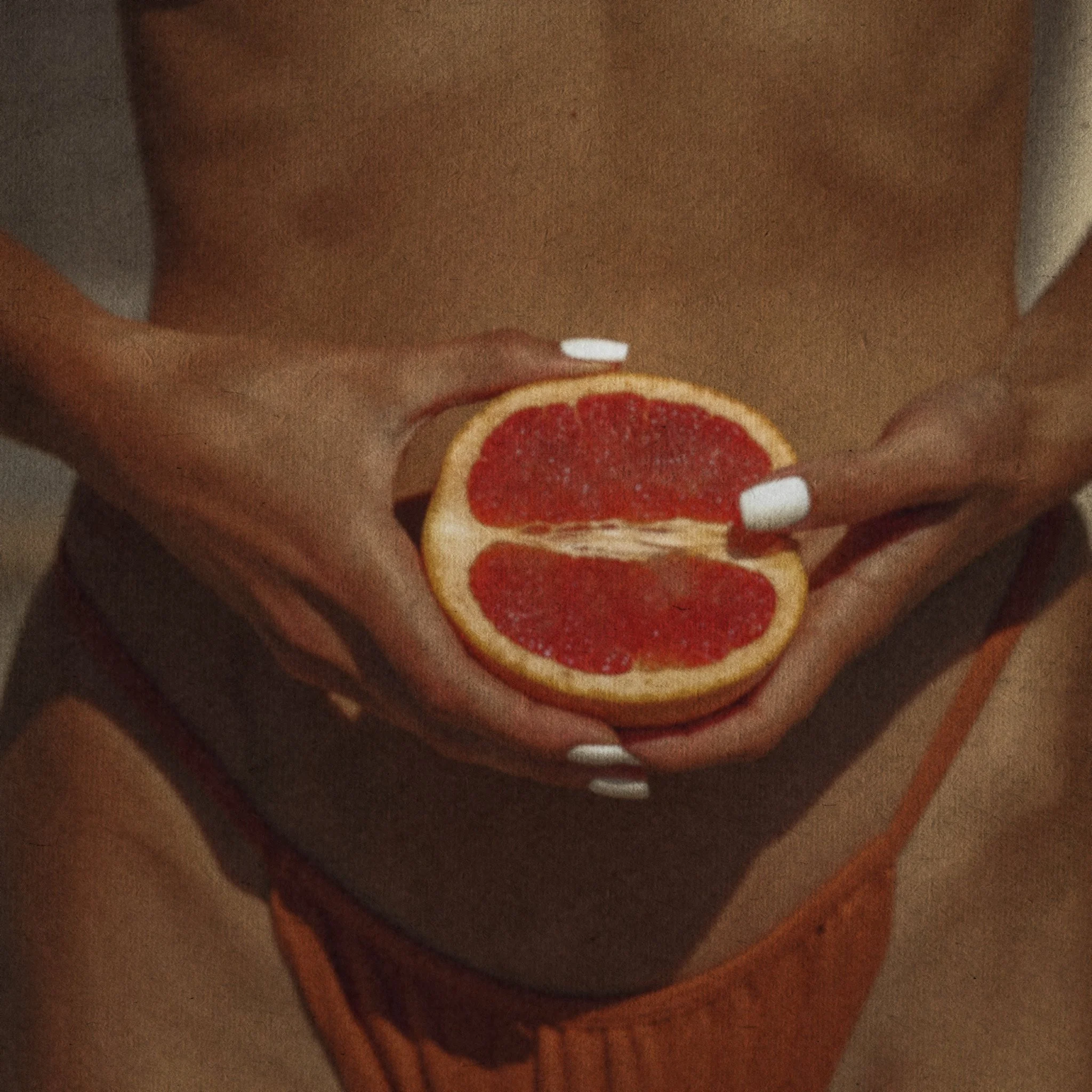 Nahaufnahme einer Person, die eine halbe Grapefruit hält, mit roten Fruchtfleisch und gelber Schale, gegen den Bauch und die Taille in einem orangefarbenen Kleid.