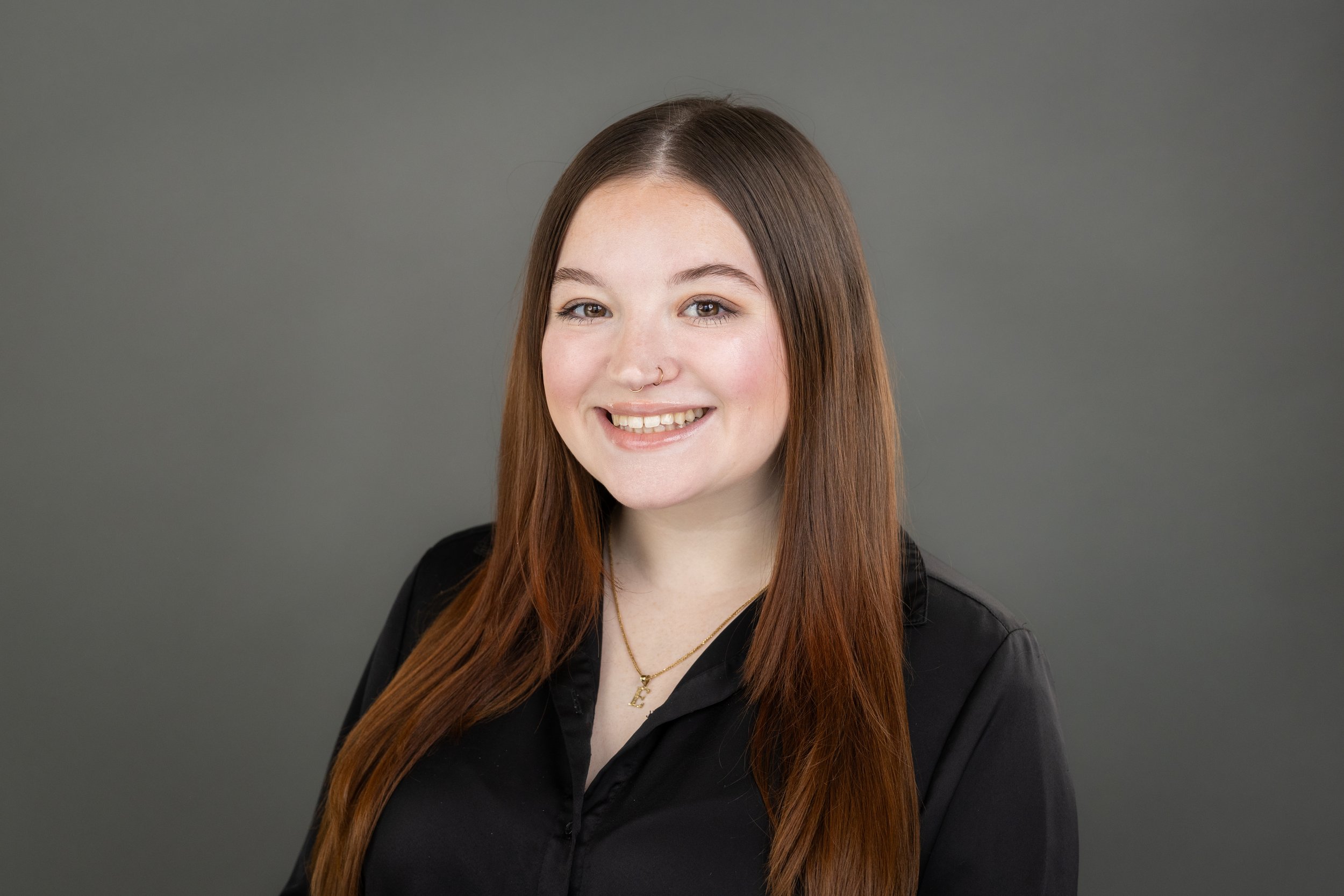 Young woman with long brown hair, smiling, wearing a black shirt, a small hoop nose ring, and a gold necklace with an 'E' pendant, against a gray background.