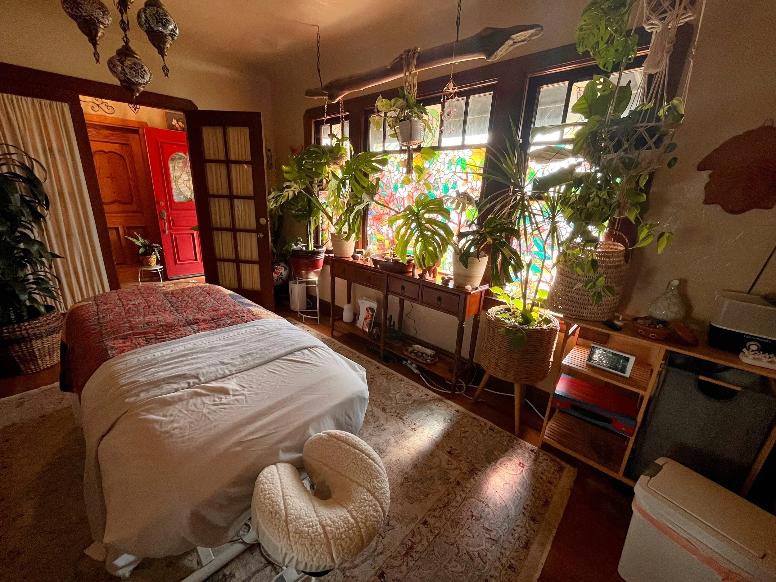 A cozy room with a massage table covered with white and red blankets, surrounded by large potted plants near a window with colorful stained glass. The room features vintage decor and warm lighting.