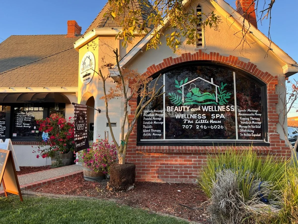 Front view of a wellness spa named 'Beauty and Wellness Wellness Spa' in a brick building with a large window displaying the business name and contact number, surrounded by plants, flowers, and a sign indicating the spa is open.