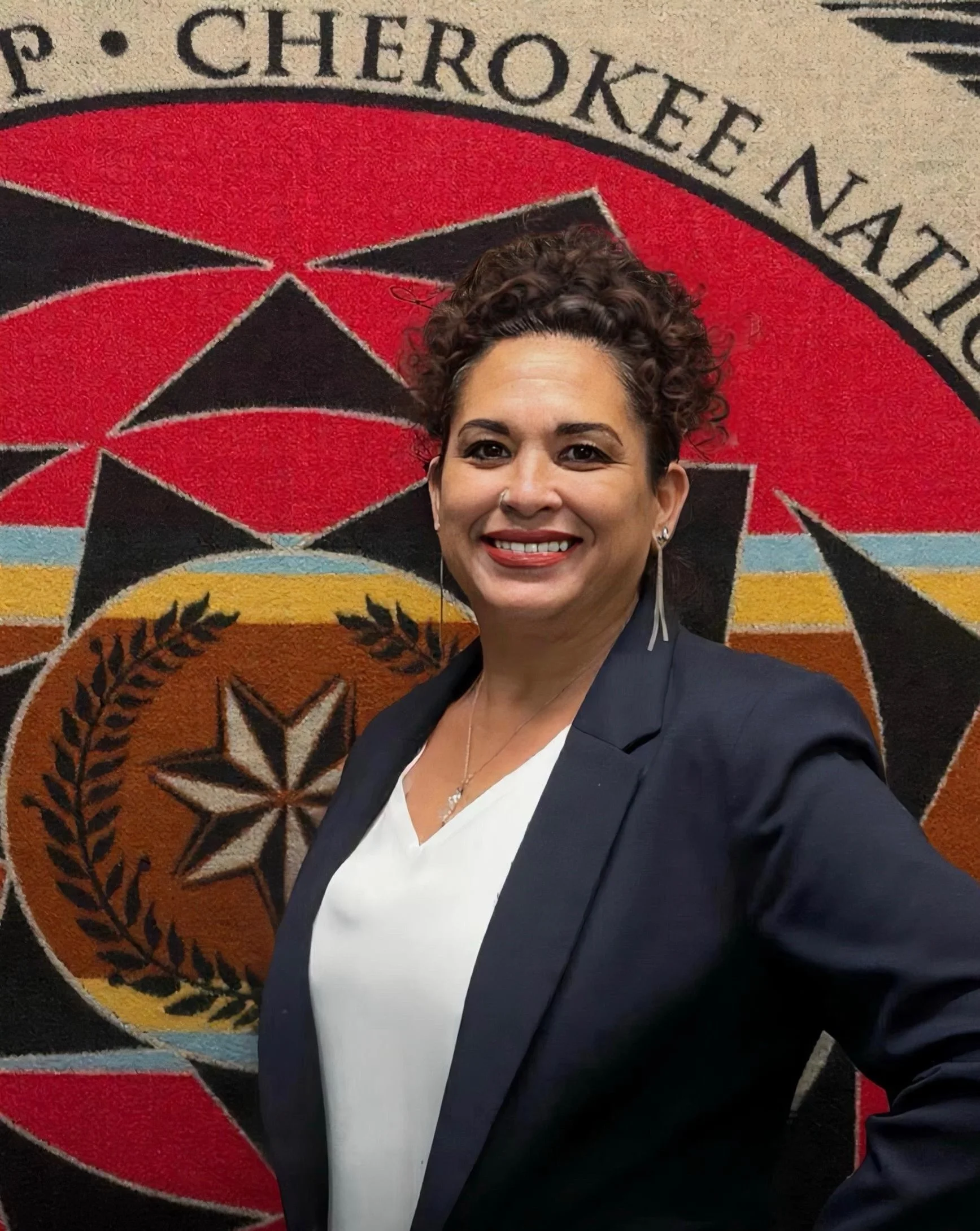 A smiling woman with short curly hair, wearing a dark blazer and white blouse, standing in front of a Cherokee Nation emblem on the wall.
