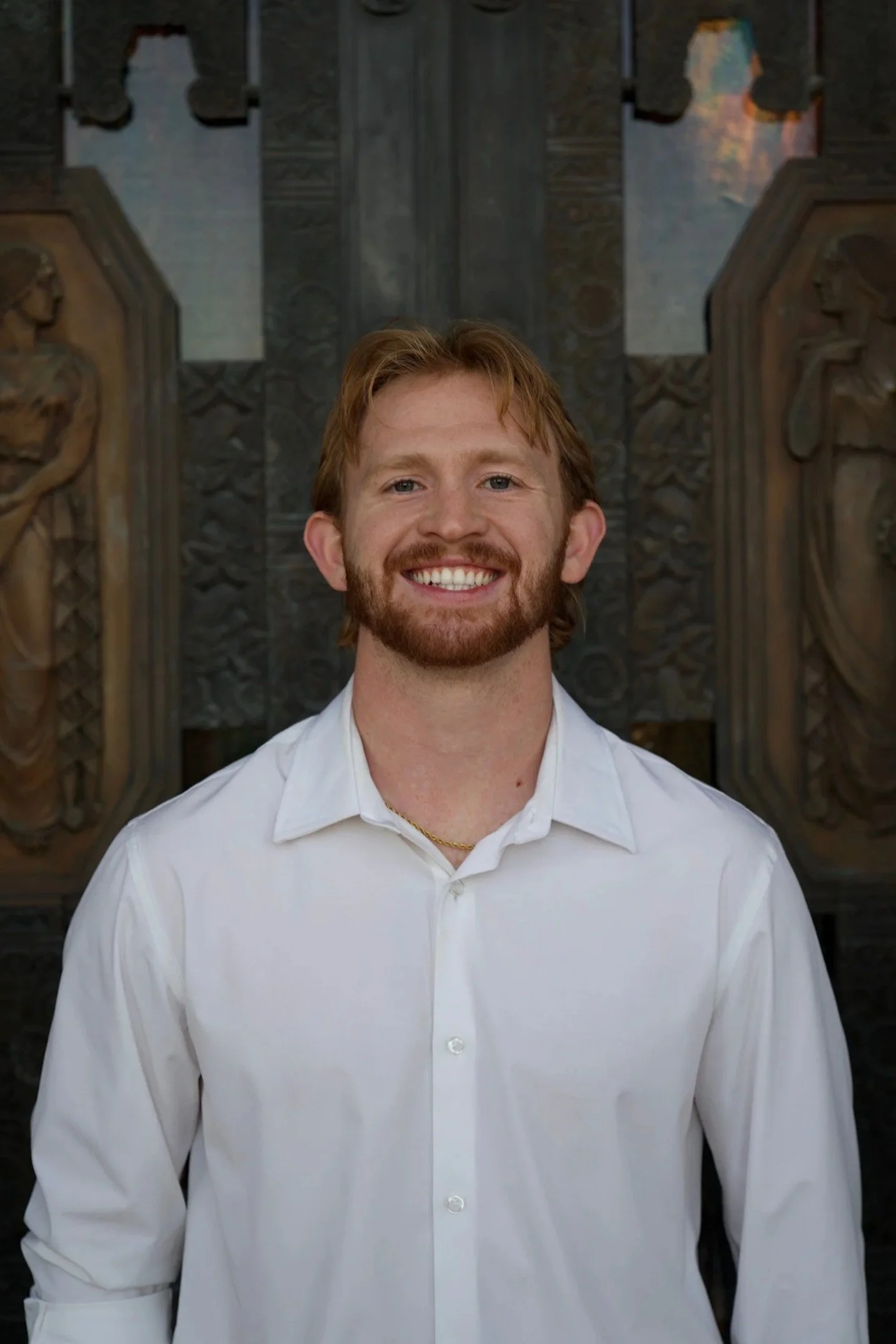 A young man with red hair and a beard, smiling in front of a dark wooden carved background, wearing a white shirt.