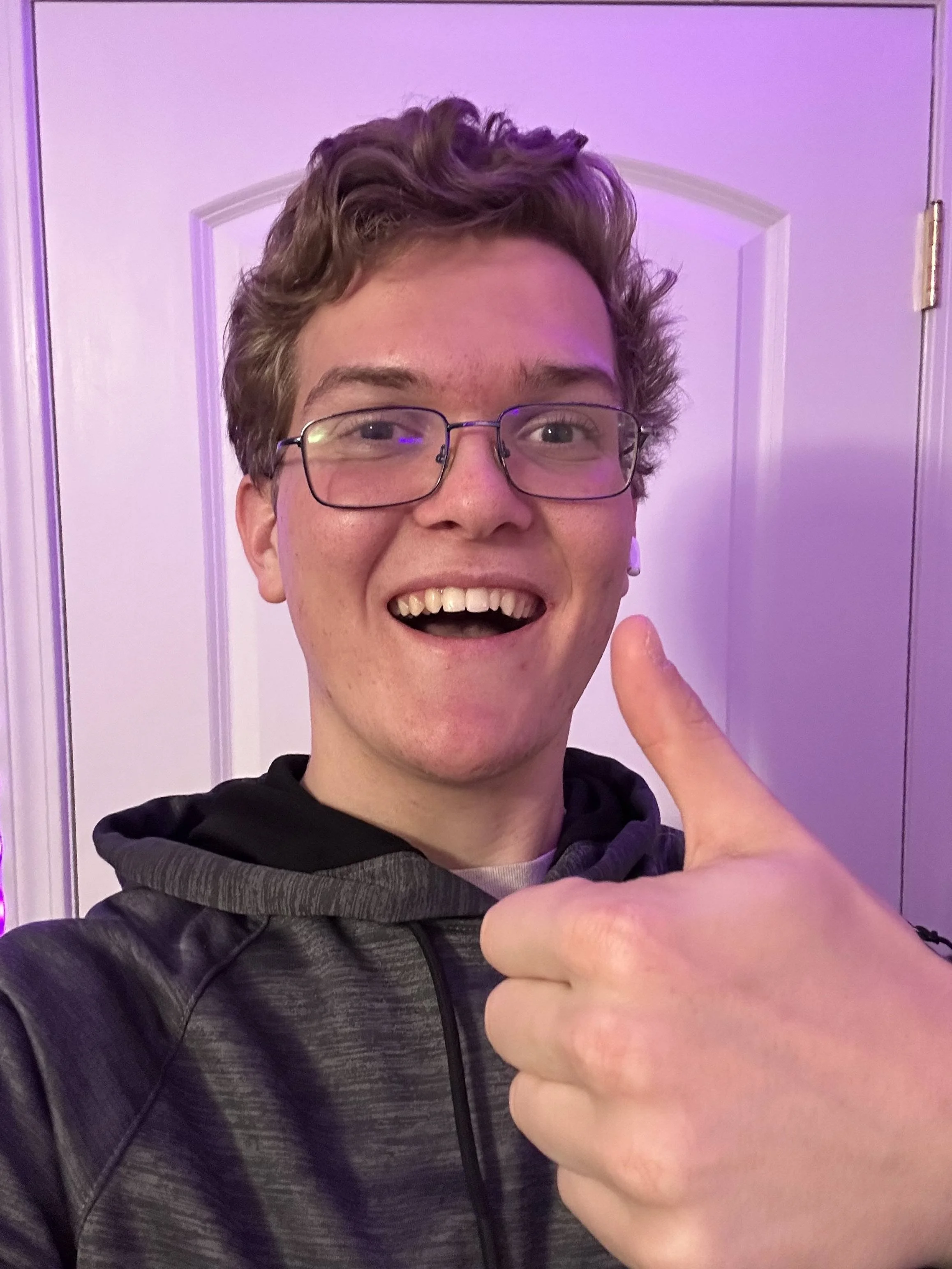 A young man with curly hair, glasses, and a wide smile, giving a thumbs-up gesture in front of a purple-lit background.