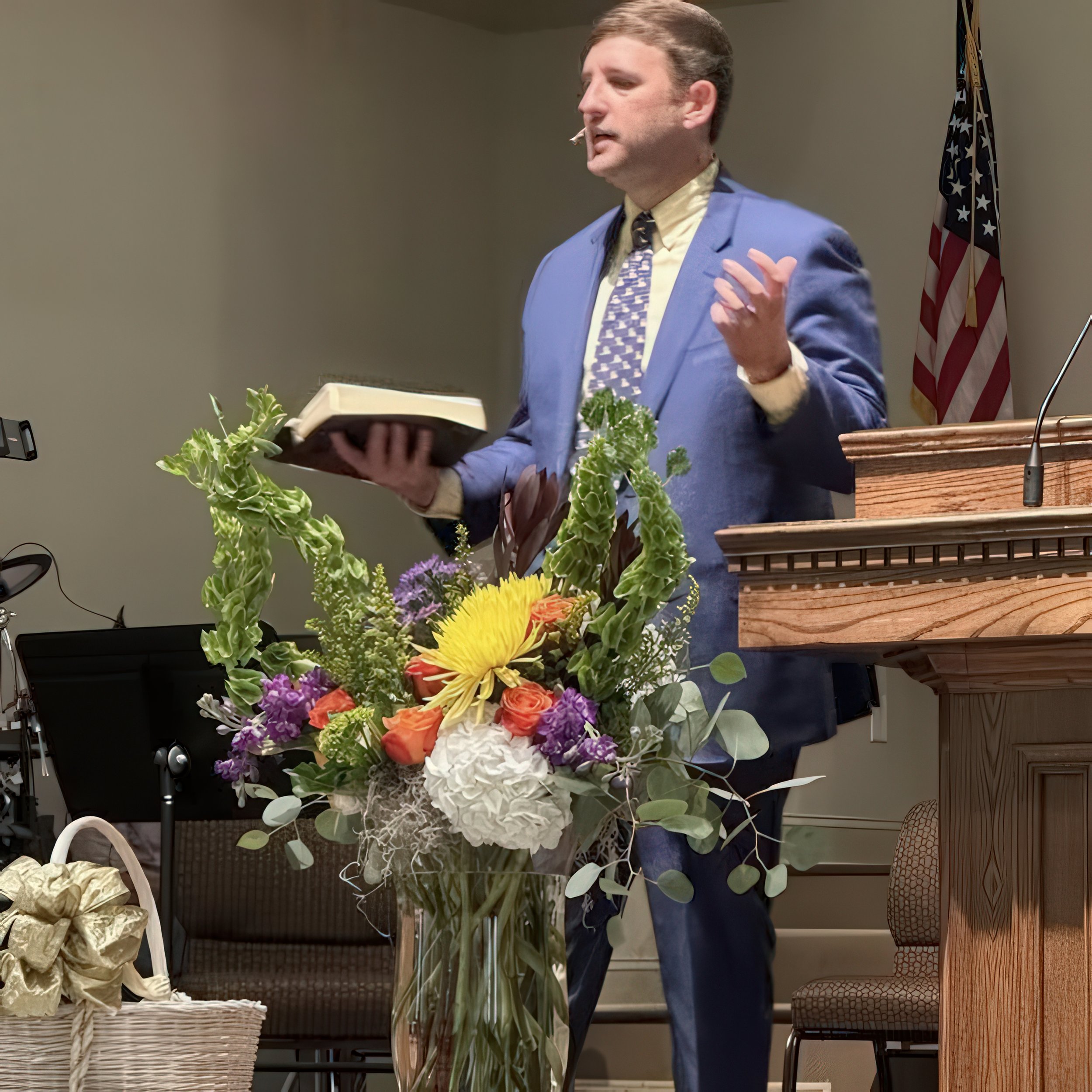 A man in a blue suit and patterned tie speaking at a podium with a flower arrangement in front. He holds a book and gesturing as he speaks, with an American flag in the background.