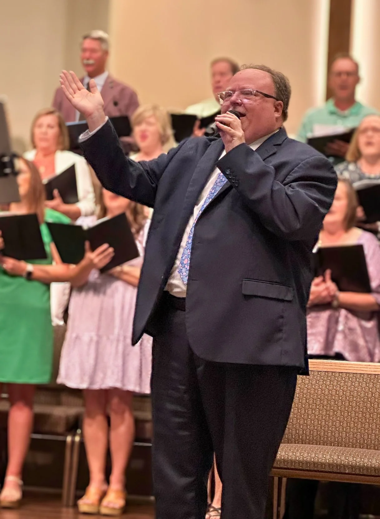 A man in a dark suit and glasses singing into a microphone, leading a choir. The choir members are holding black songbooks and appear to be singing, with some smiling. The background is blurred with warm lighting.