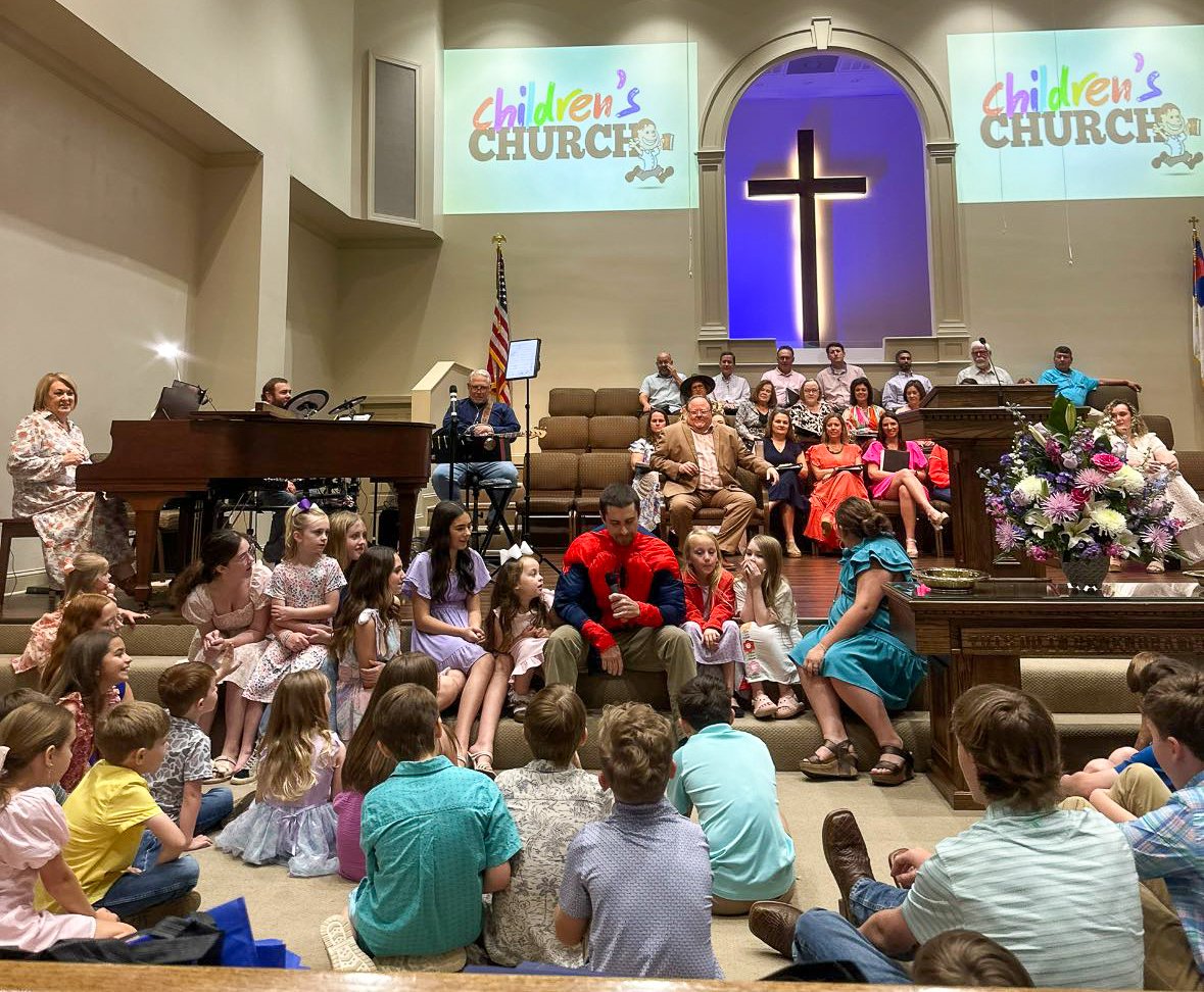 Children's church service in a church with a large cross at the back and a priest or pastor speaking to children sitting on the floor. Adults are seated on chairs behind them, and children are on the stage and sitting on the floor. There are two large screens with the children's church logo.