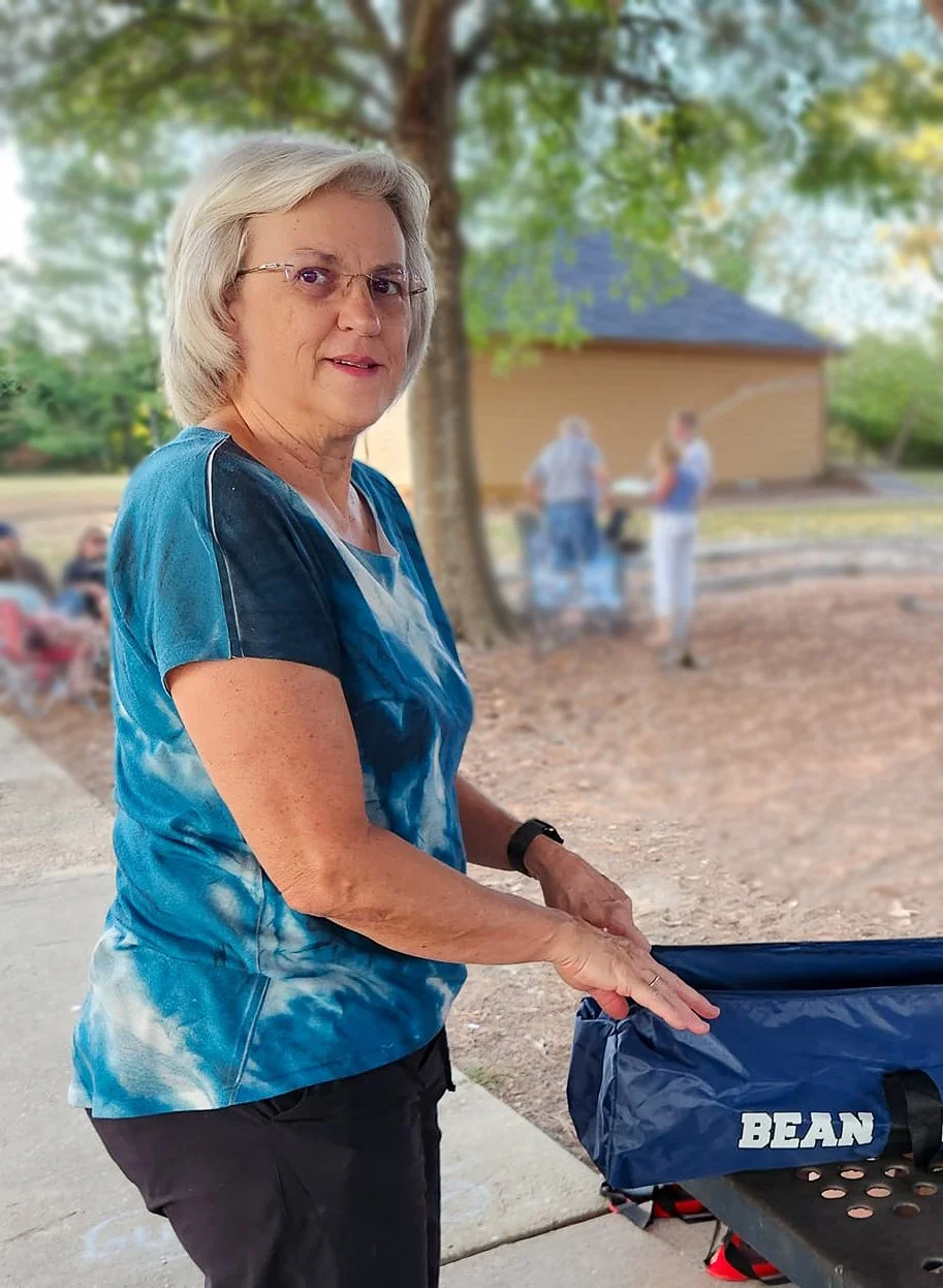 Older woman with blonde hair, glasses, wearing a blue tie-dye shirt and black pants, standing outdoors near a picnic table, with people and a building in the background.