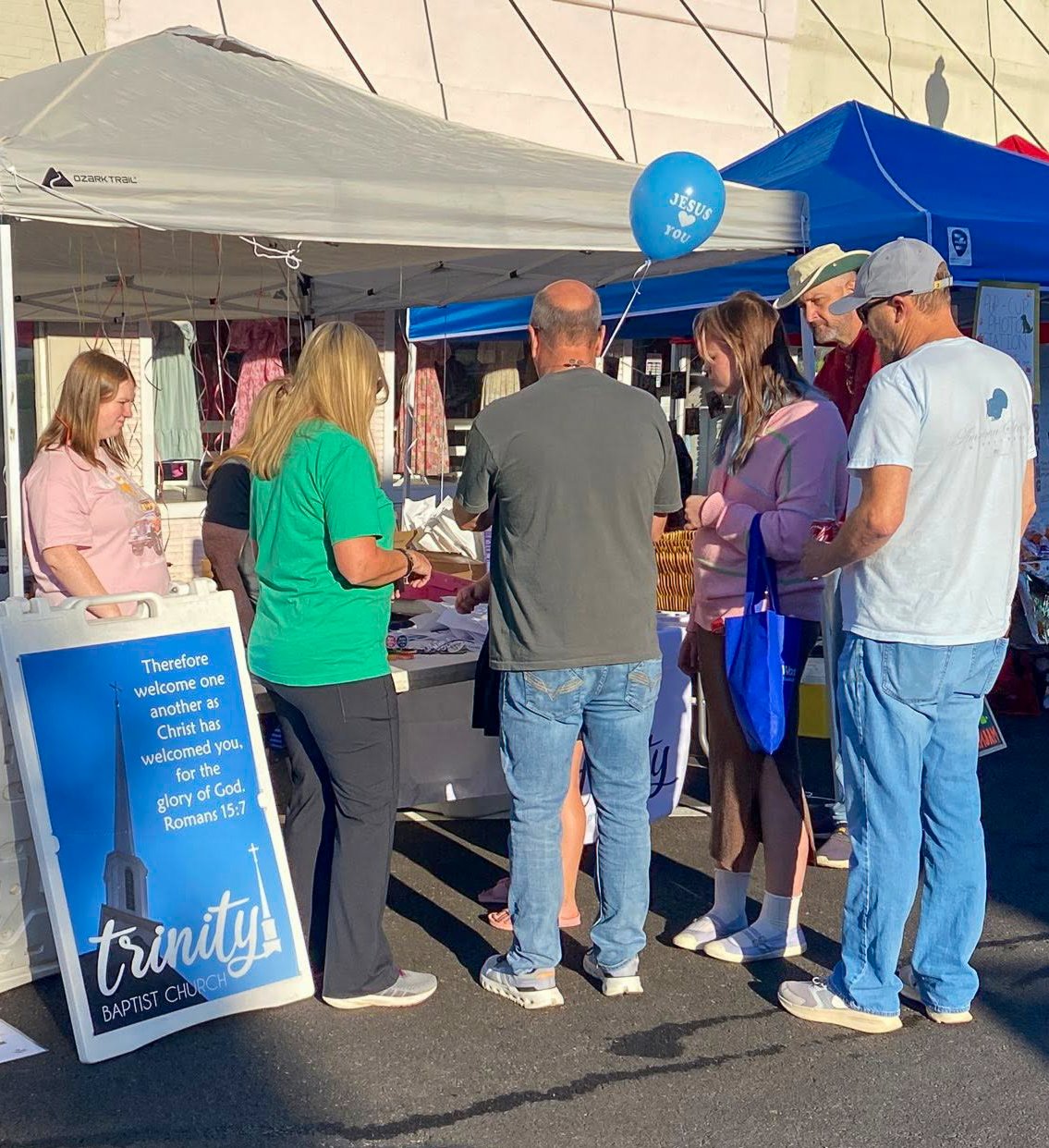 People gathered at a Trinity Baptist Church booth during an outdoor event, with a sign displaying a Bible verse and a blue balloon reading 'Jesus you'.