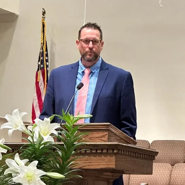 A man in glasses, wearing a blue suit, light blue shirt, and pink tie, stands behind a wooden podium with white lilies in the foreground. An American flag is in the background.