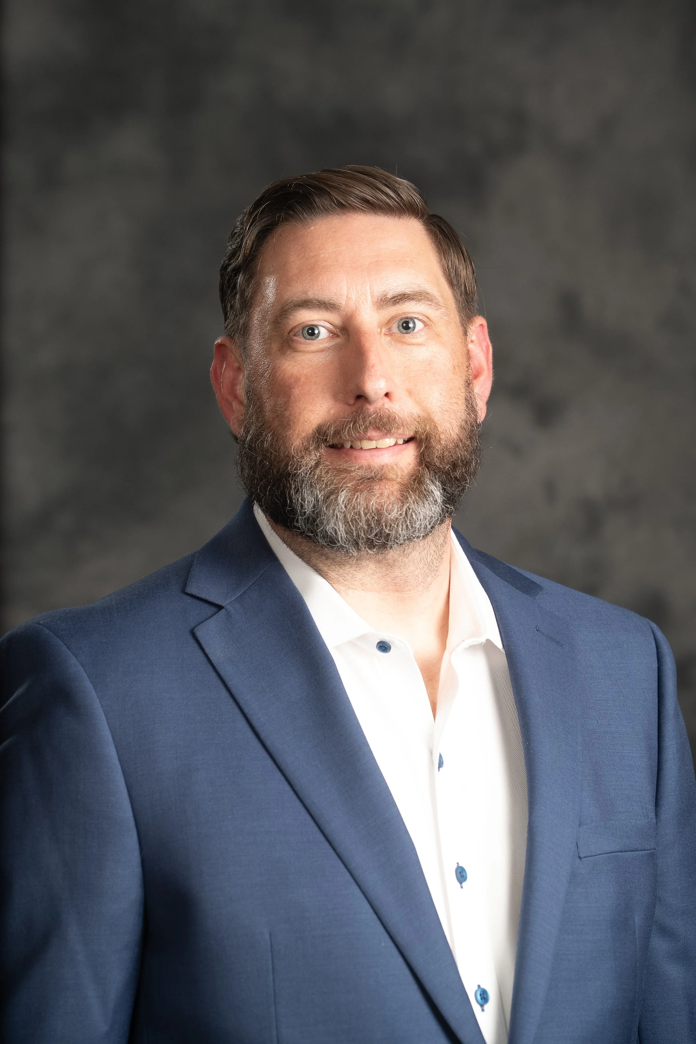 A man with dark hair, light skin, and a beard wearing a blue suit jacket over a white collared shirt, smiling against a dark, blurred background.