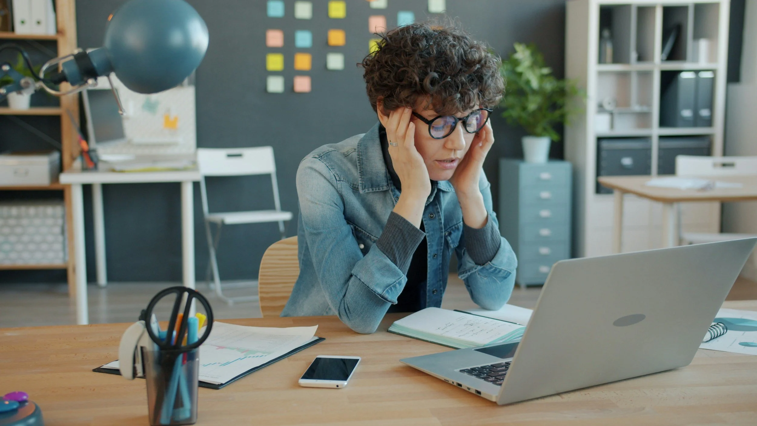 A woman with curly hair and glasses sitting at a desk in an office, looking at her laptop with a stressed expression, holding her head with her hands. There are notebooks, a smartphone, and a pen holder on the desk, with a bookshelf and a gray filing cabinet in the background.