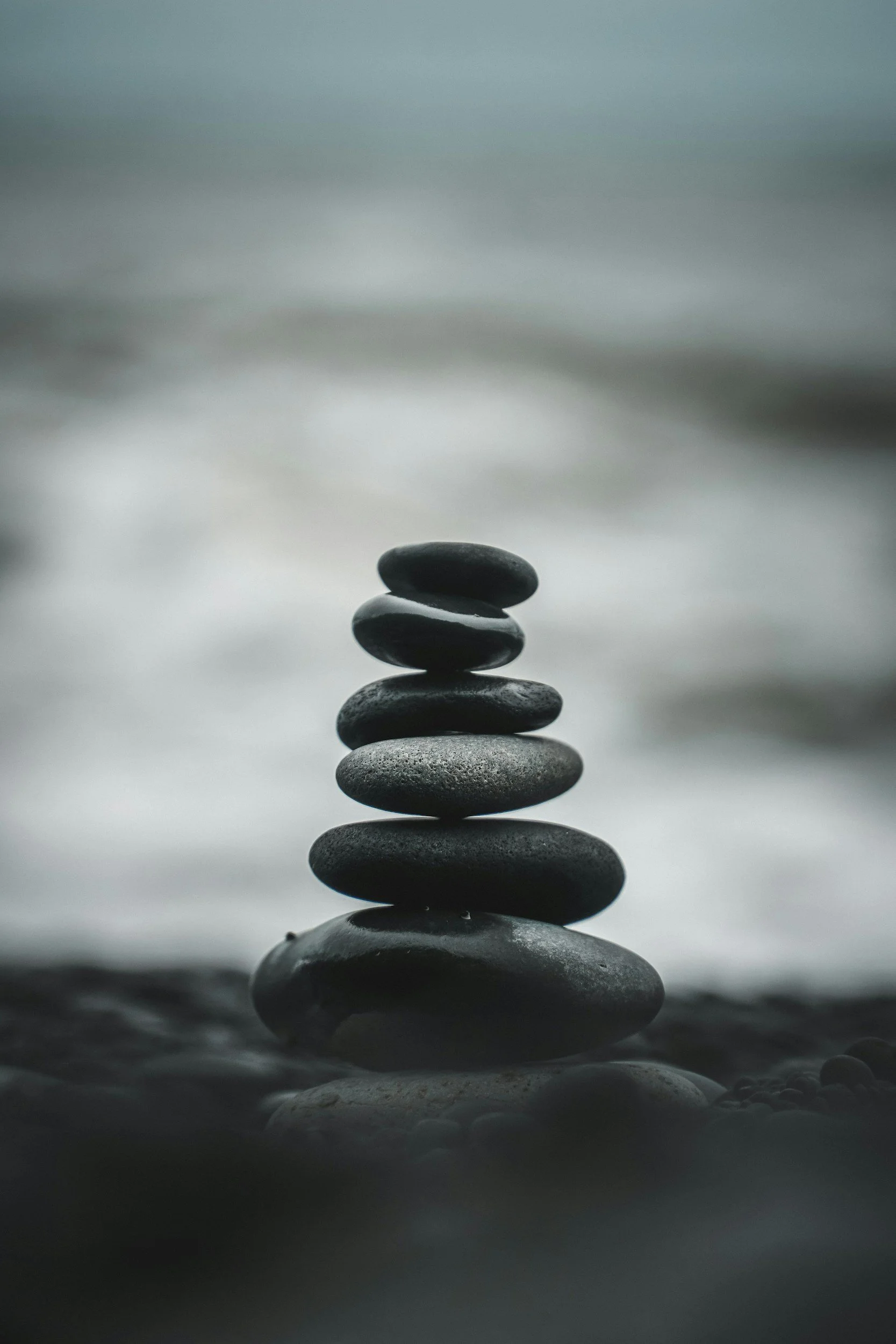 A stack of smooth, black stones balanced on top of each other on a wet surface near a body of water, with a blurred background.