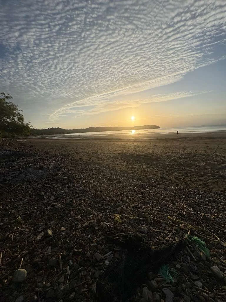 Sunset over a beach with scattered rocks and a person walking along the shoreline, with clouds in the sky.