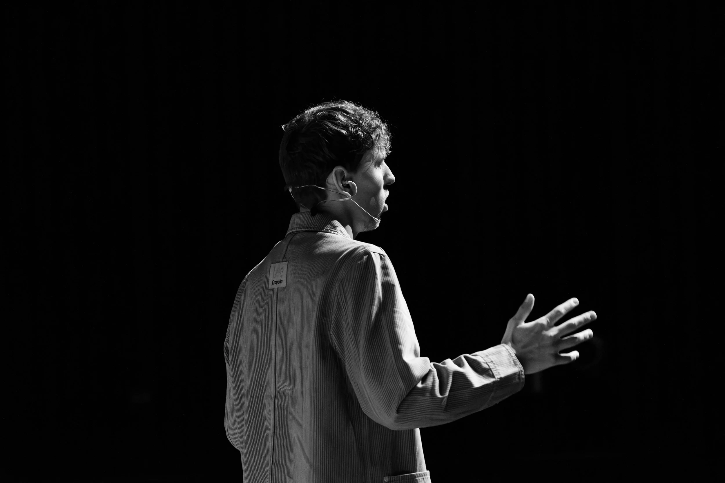 A black-and-white photo of a person wearing a headset microphone, facing sideways with their hand raised, on a dark background.