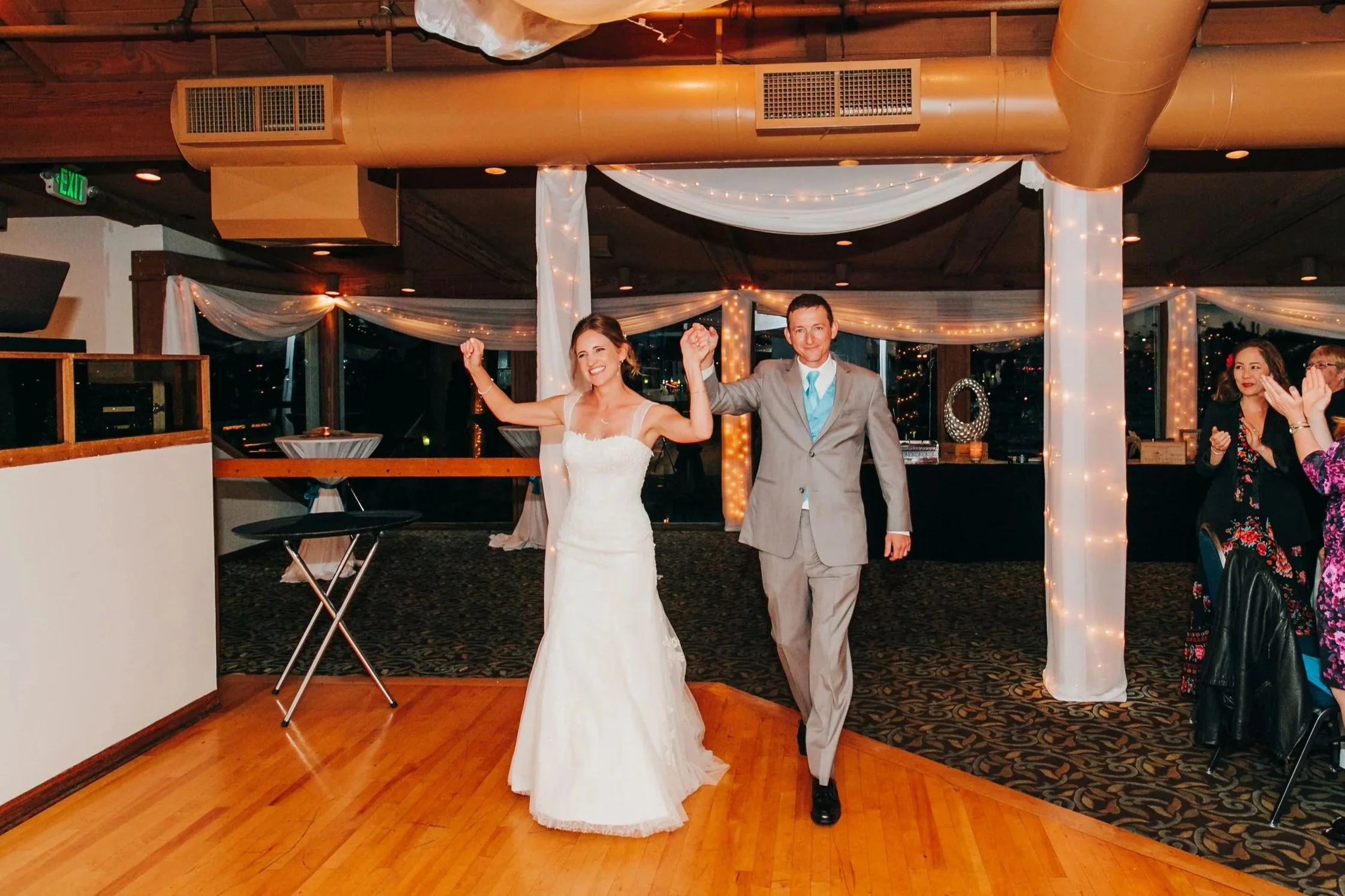 A bride and groom holding hands and smiling at their wedding reception, dancing together in a decorated hall with fairy lights and draped fabric.