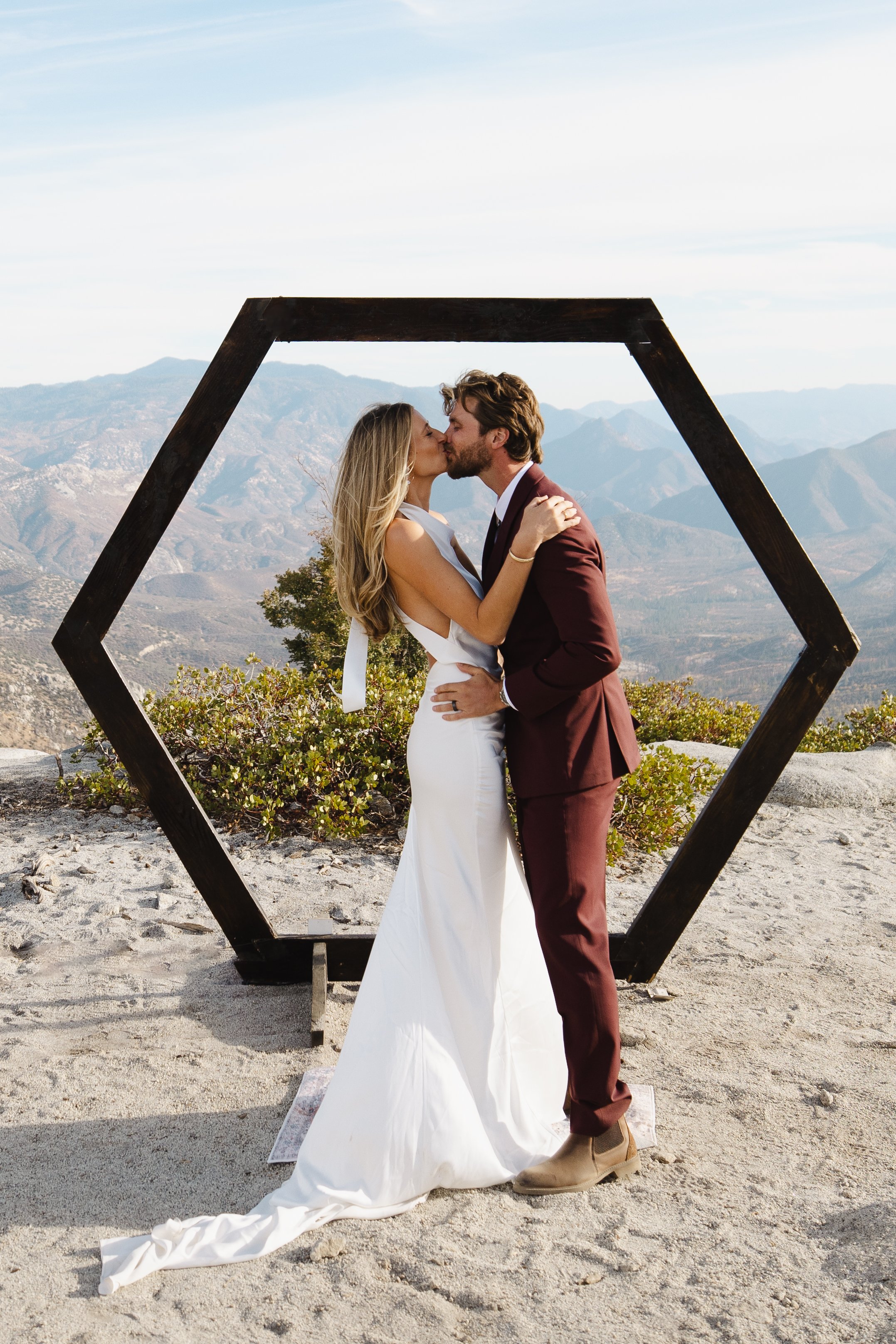 A couple kissing at a scenic outdoor location with mountains in the background, framed by a large hexagonal wooden structure.
