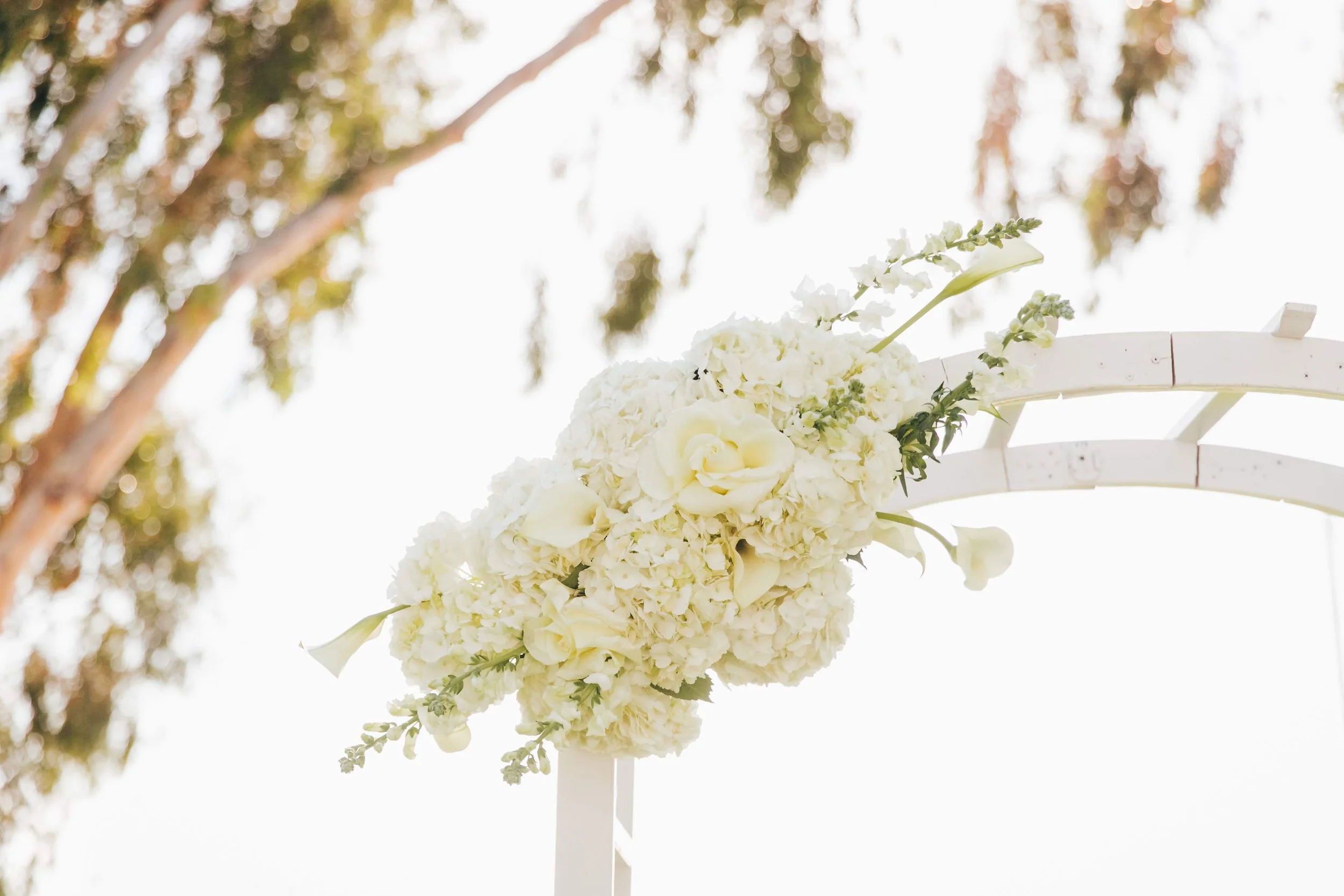 White floral arrangement attached to an arch, with a background of blurred trees and bright sky.