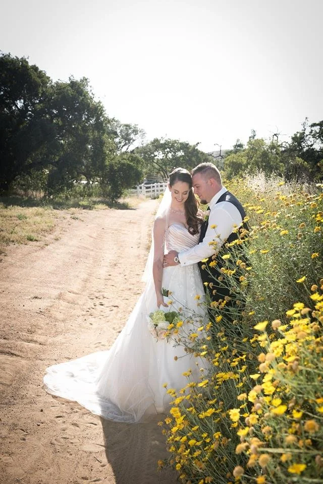 Bride and groom smiling together on a dirt path surrounded by yellow wildflowers and trees during daytime.