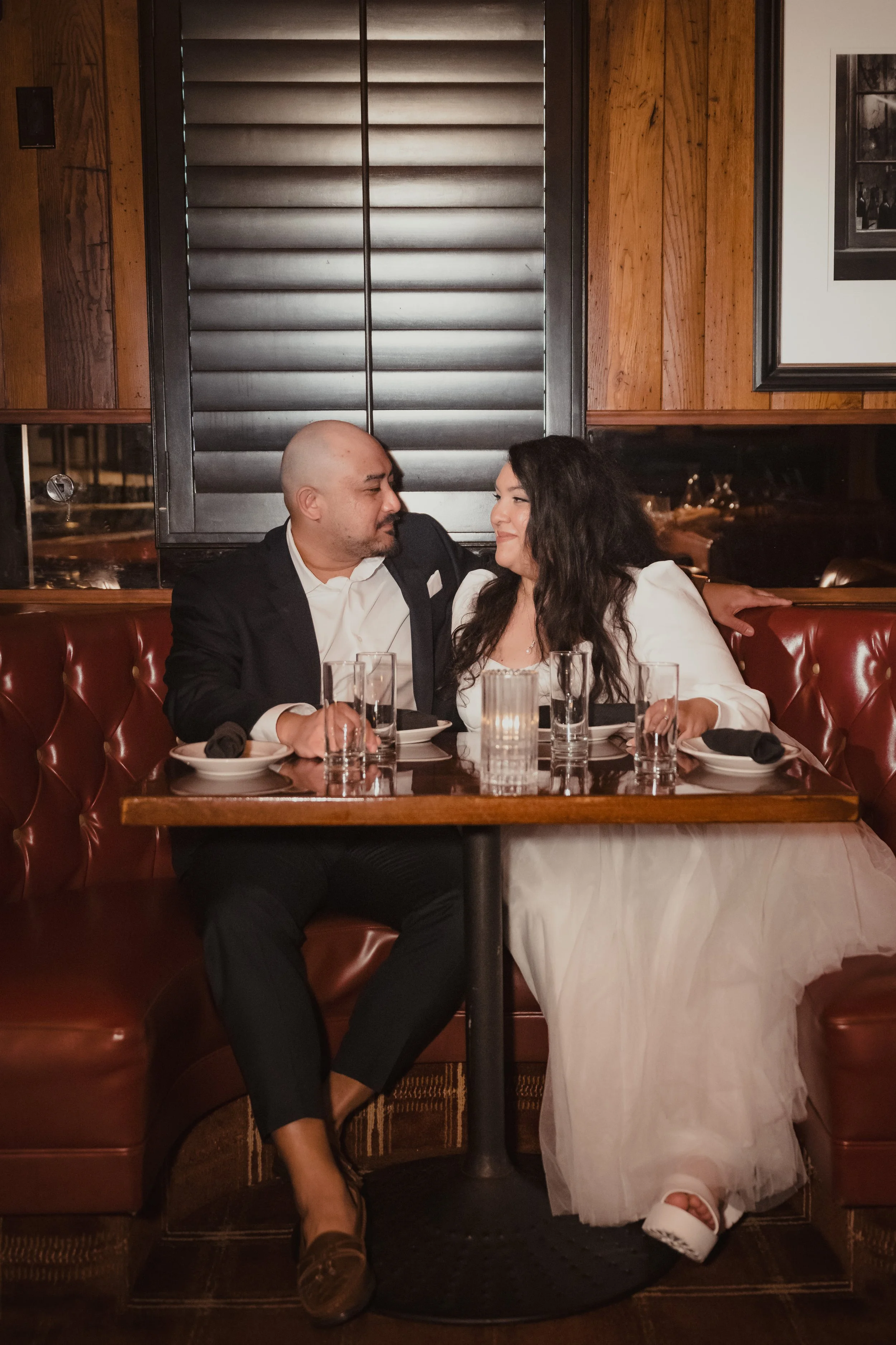A couple sitting at a restaurant table, gazing at each other, with a warm and intimate atmosphere, candles on the table, and wooden walls in the background.