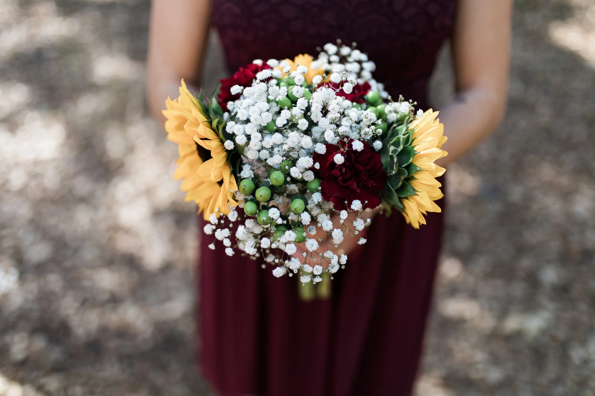 Person in a maroon dress holding a bouquet of yellow sunflowers, white baby's breath, red carnations, and green berries, with a blurred outdoor background.