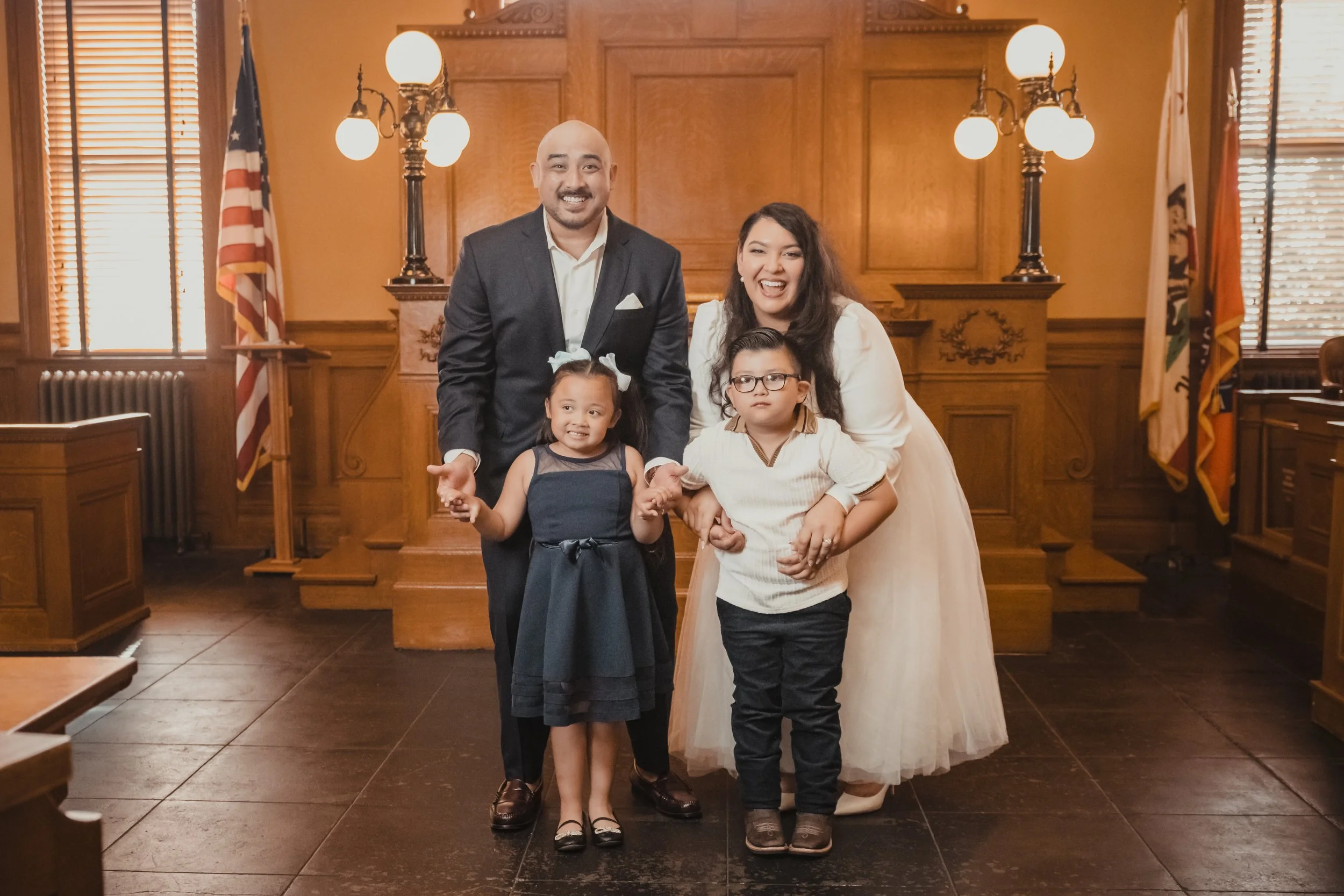 A family of four, including a man in a suit, a woman in a white dress, and two children, posing and smiling inside a wooden courtroom with flags and lighting fixtures.