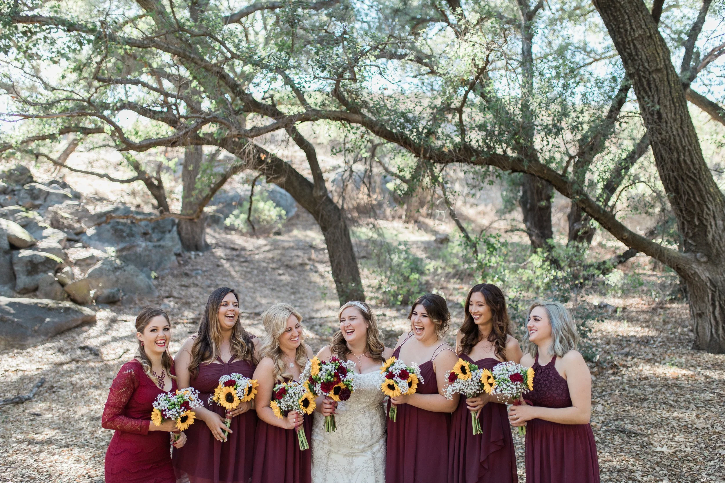 Group of seven women, including a bride in white and bridesmaids in burgundy dresses, standing outdoors under trees, holding colorful bouquets, smiling and enjoying a wedding celebration.