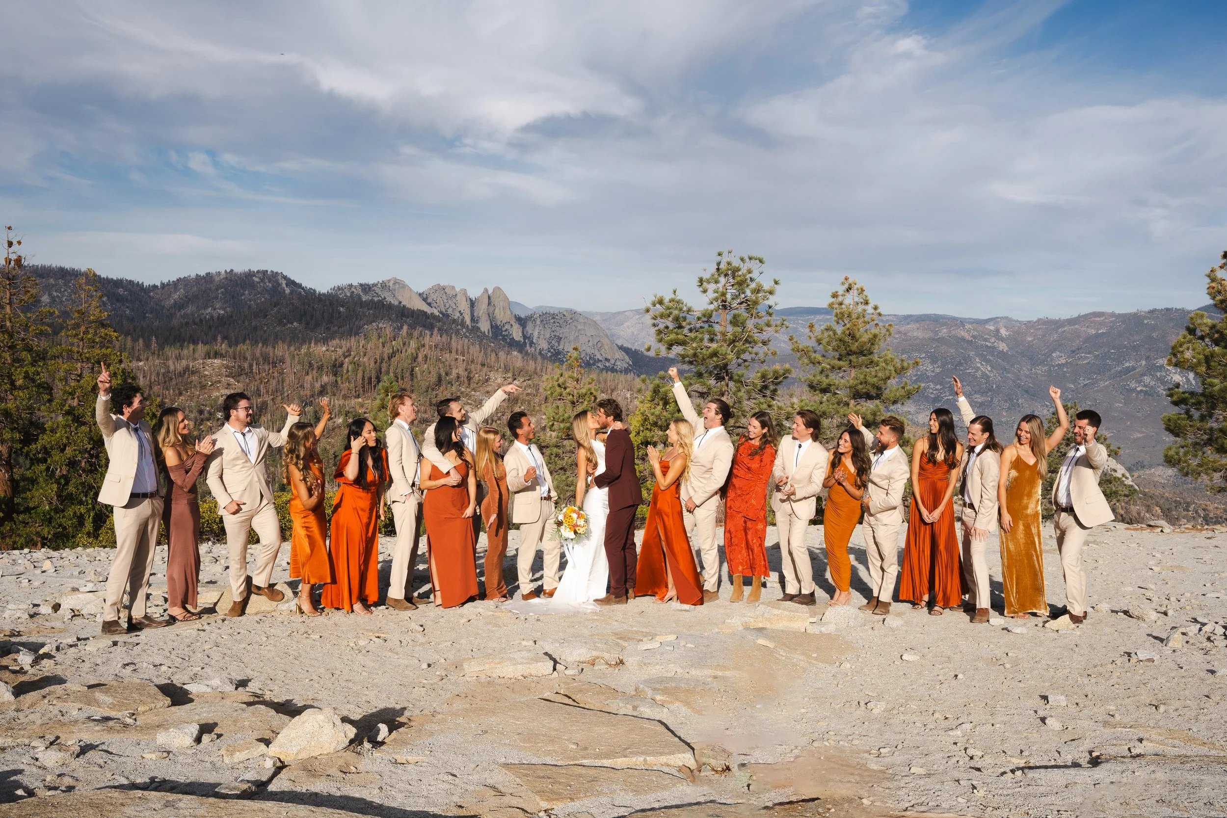 A wedding party standing outdoors on rocky terrain with mountains and trees in the background, celebrating and raising their hands.