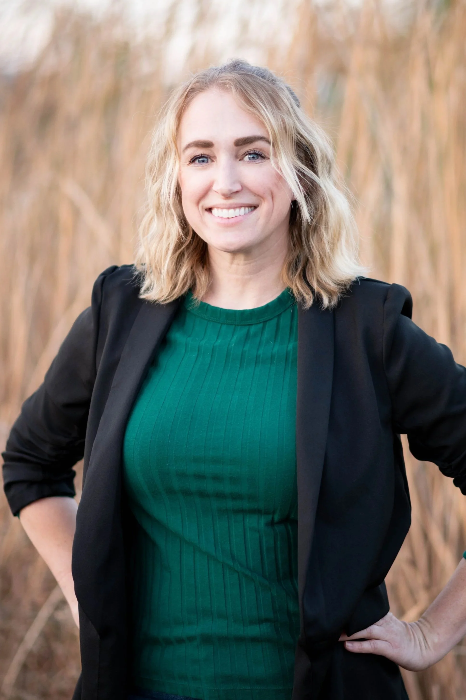A woman with wavy blond hair smiling outdoors, wearing a green shirt and black blazer, standing amidst dry tall grass.